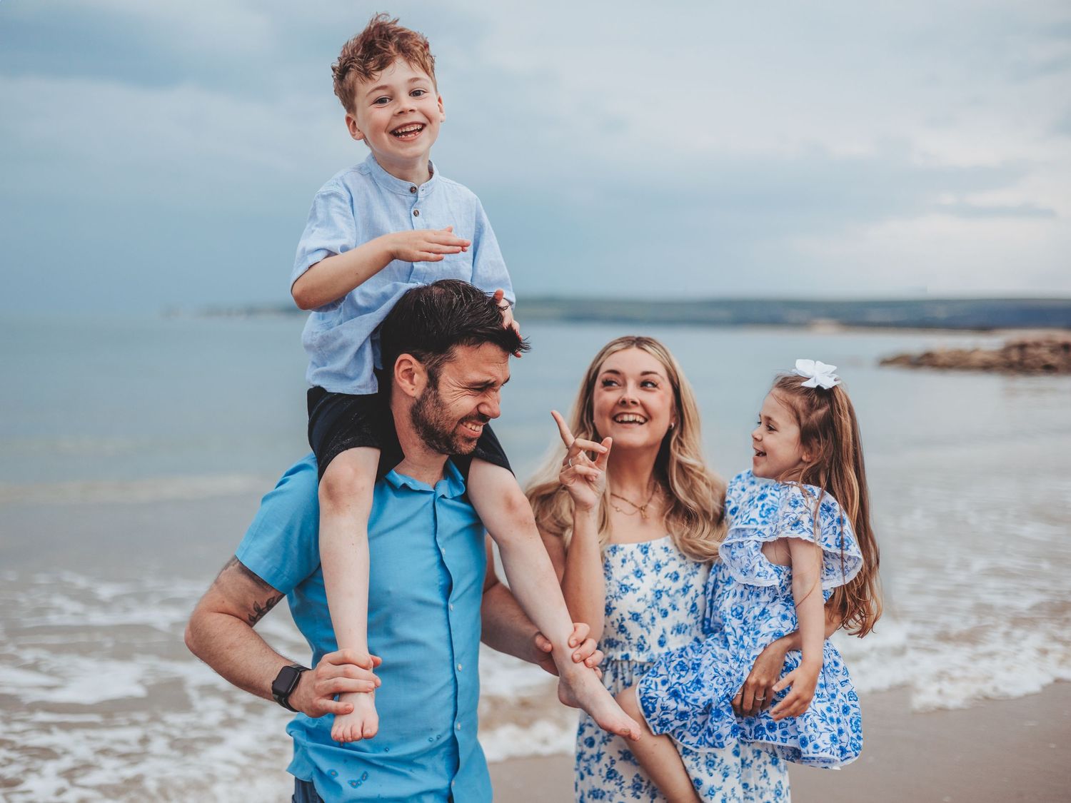 Happy family of four enjoying a beach day, parents carrying young children, smiling against a cloudy seaside backdrop.