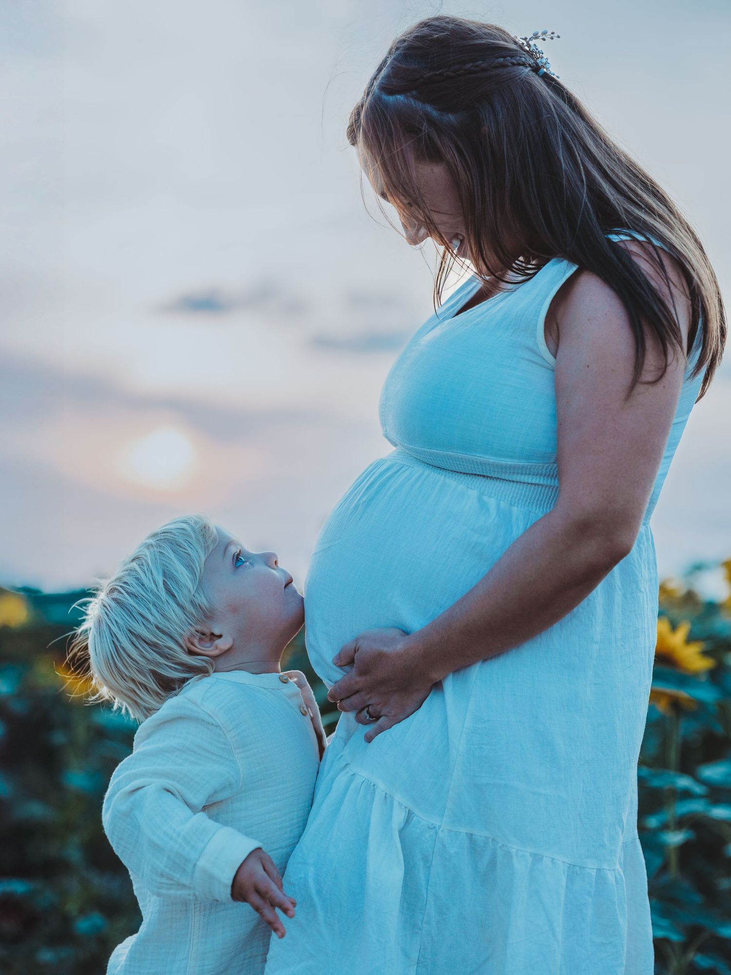 A touching moment at sunset shows a young child looking up at a pregnant woman in a turquoise dress standing in a flower field.