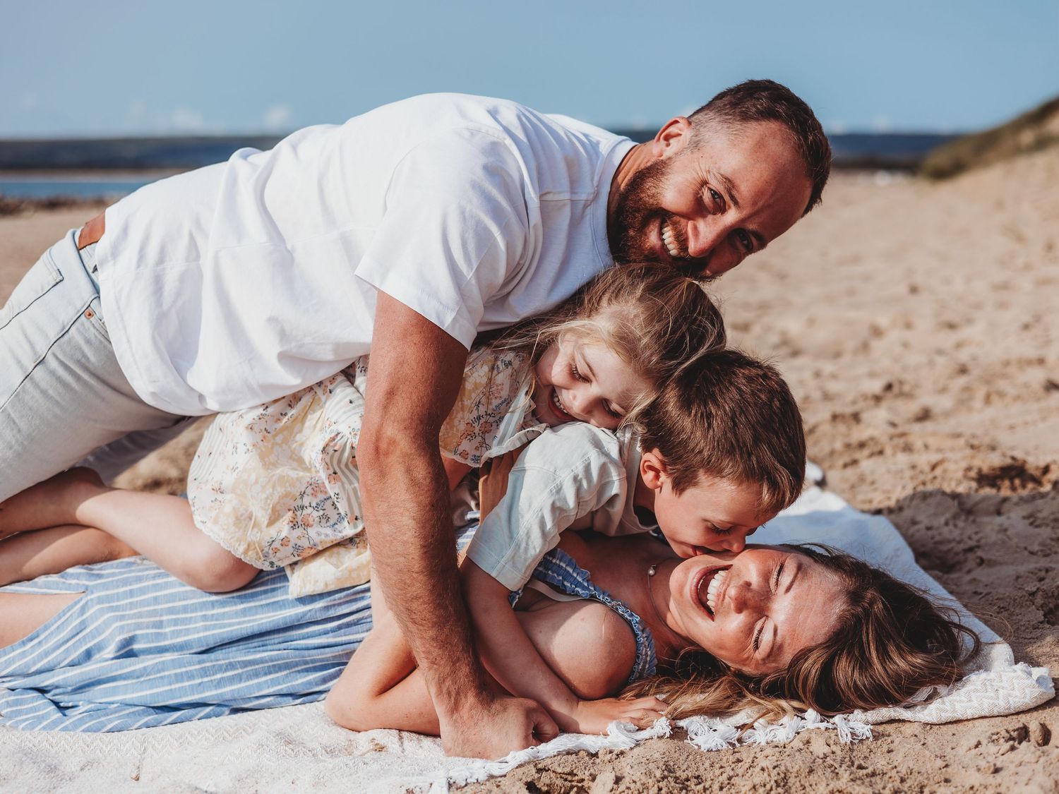 A loving family enjoys playful moments together while lying on a sandy beach in white clothing.