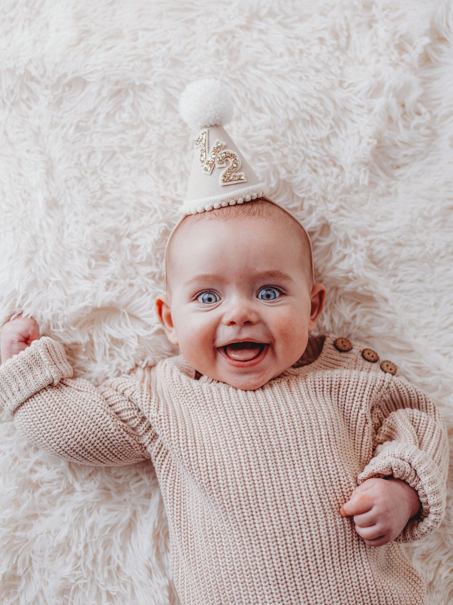 A smiling baby wearing a knitted beige sweater and white winter hat lying on a fluffy white blanket.