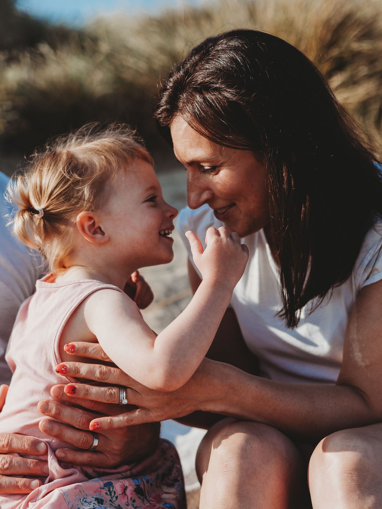 A smiling mother and toddler girl share a joyful nose-to-nose moment on a sunny beach day.