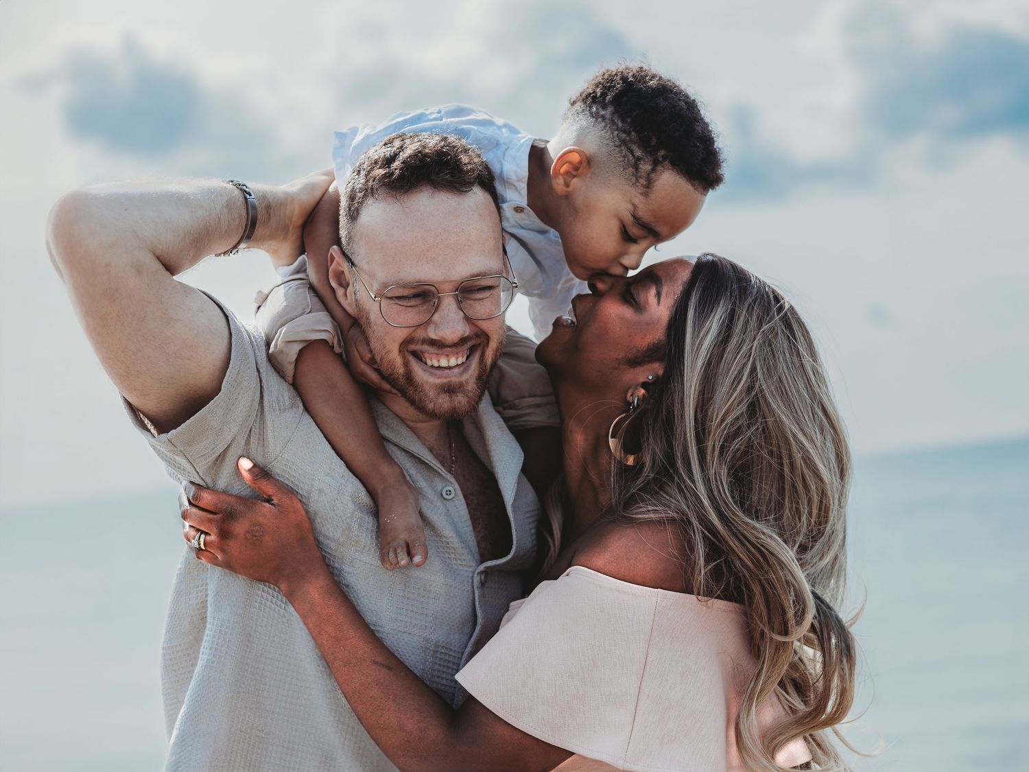 Happy family at the beach, father carrying child on shoulders while mother kisses baby, smiling outdoors.