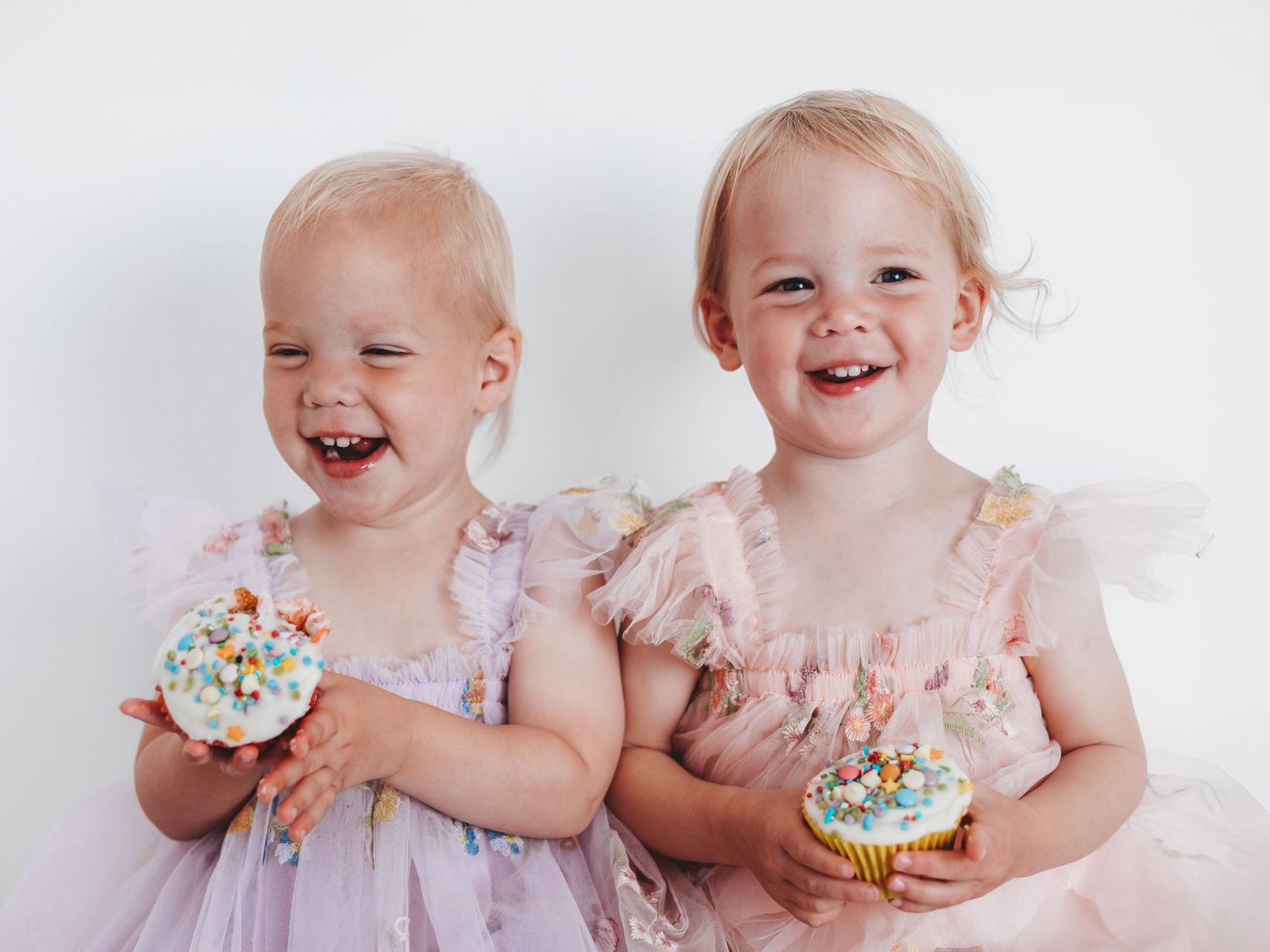 Two toddler girls in pink dresses holding colorful cupcakes, smiling joyfully against a white background.