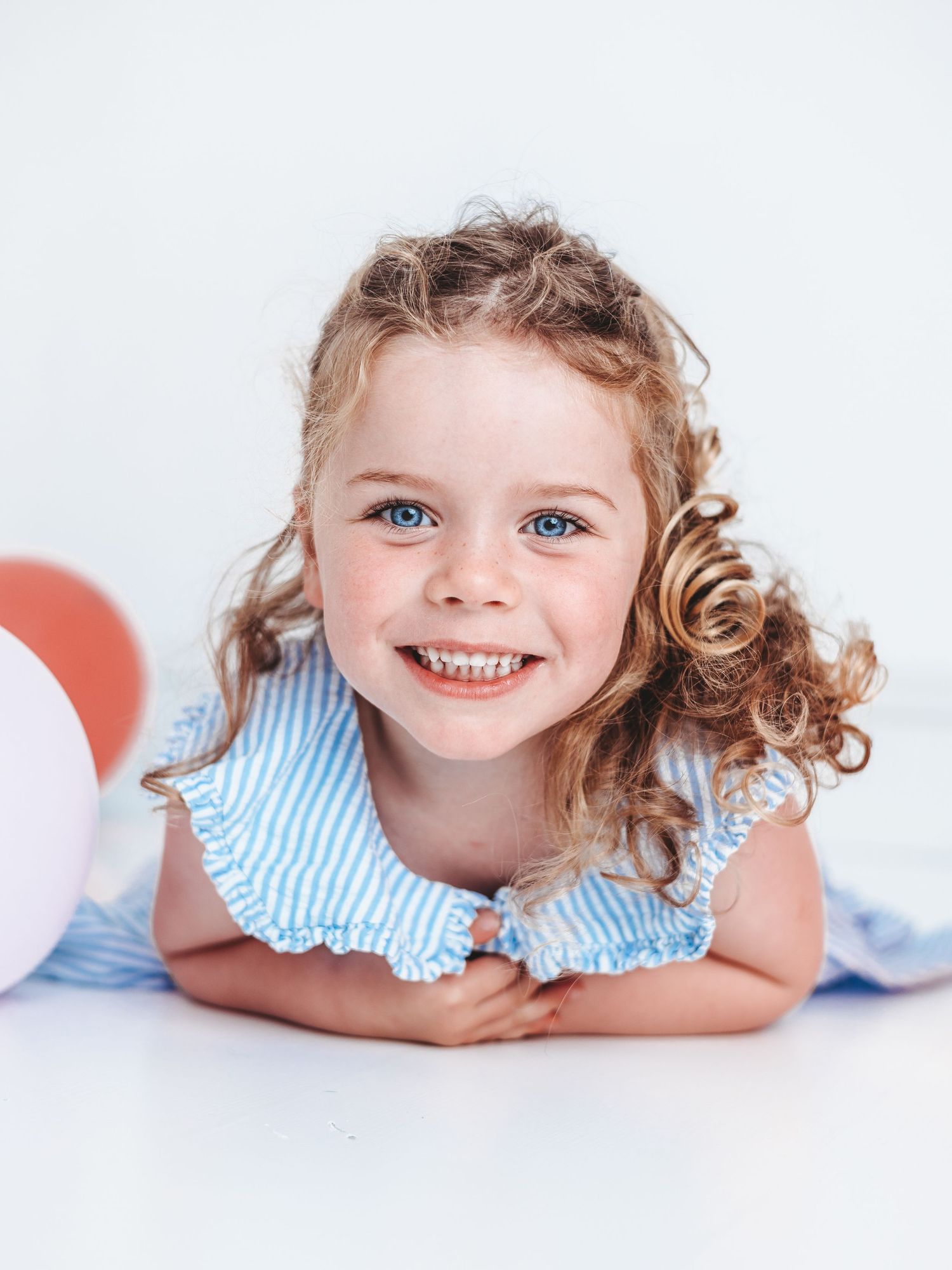 A smiling young girl with blue eyes and curly hair wearing a blue striped dress, lying on the floor with balloons.