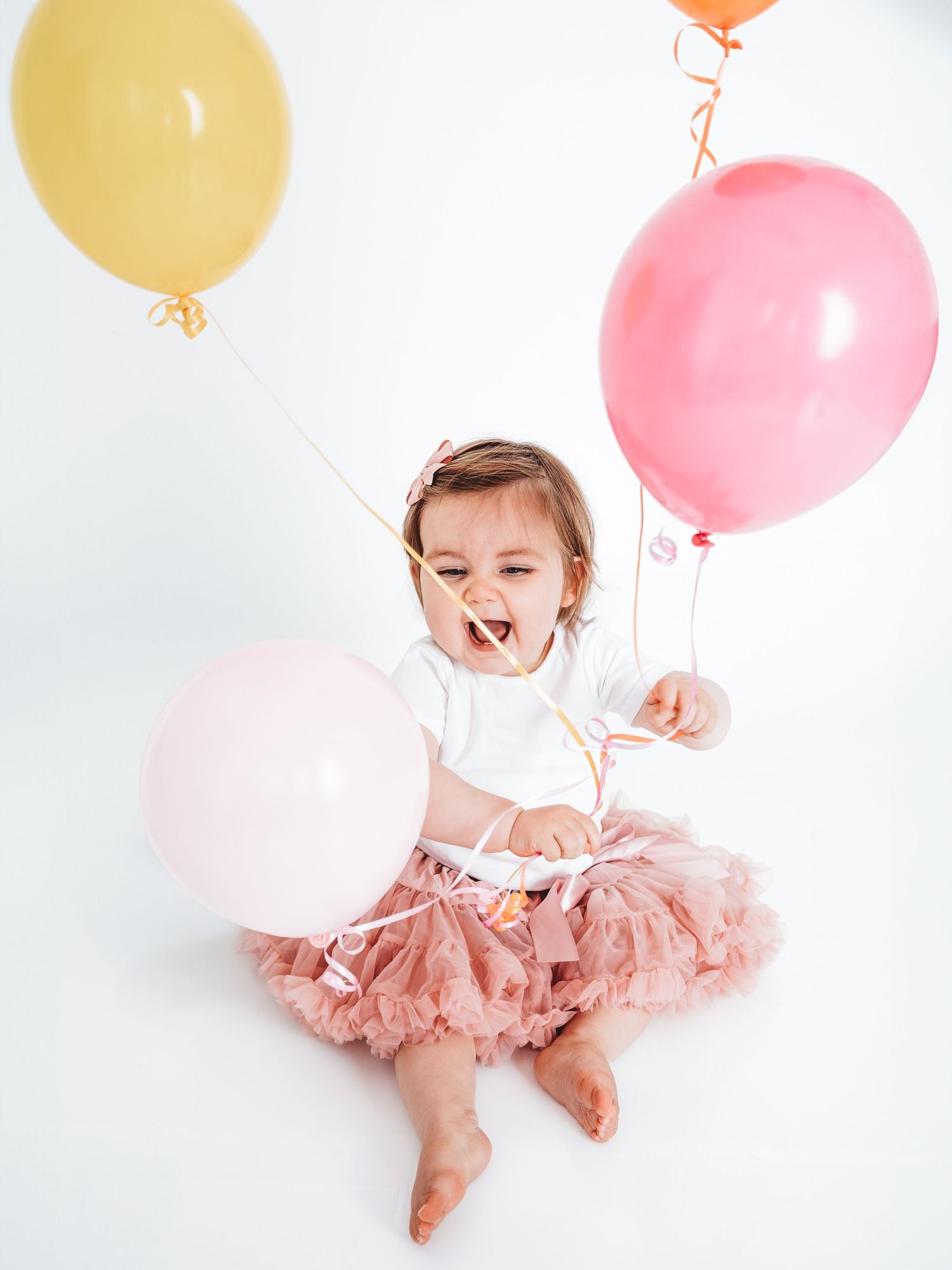 Happy toddler girl in pink tutu skirt sitting on white floor holding colorful balloons at birthday party.