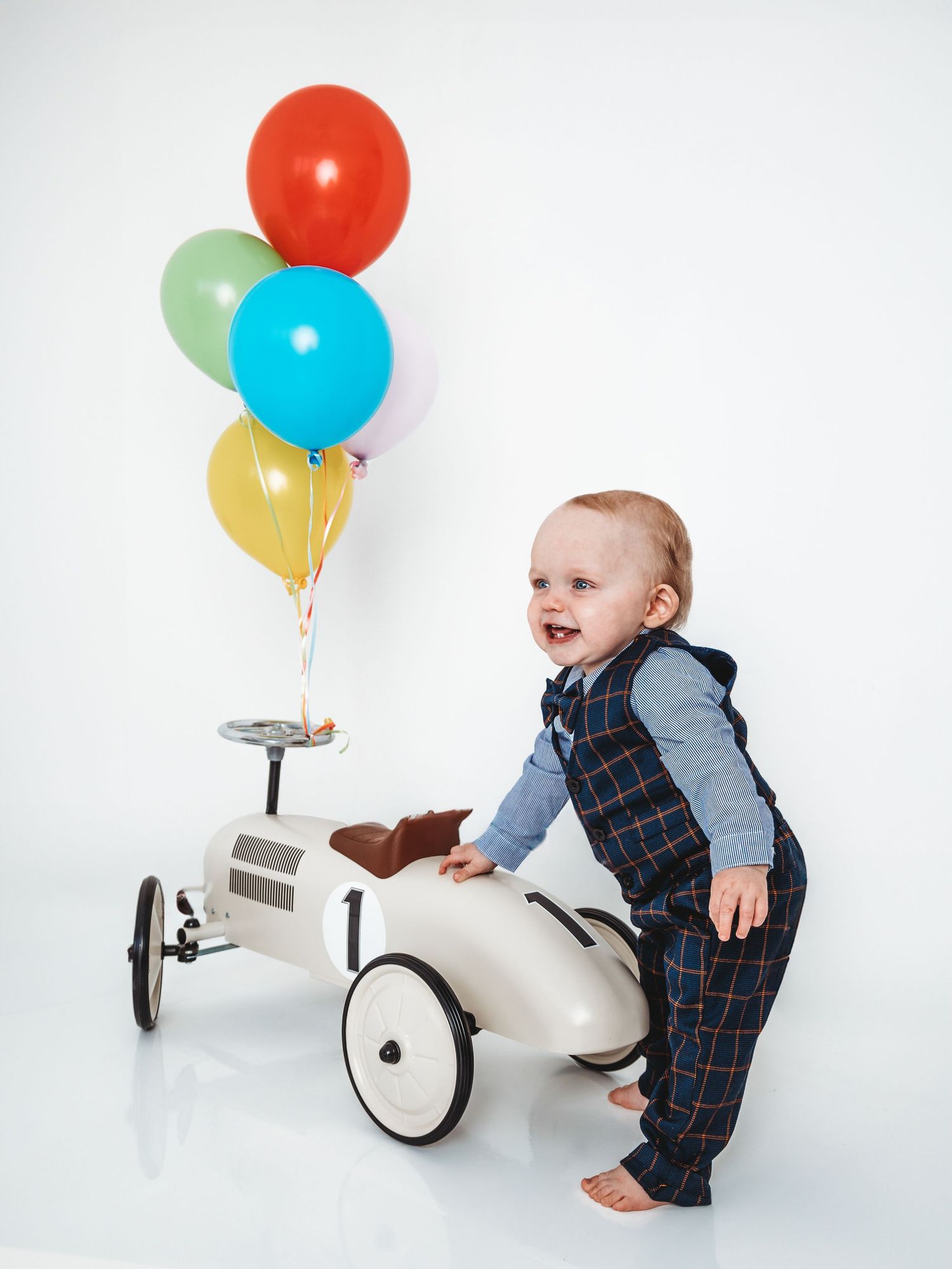 Smiling baby boy in plaid vest sits on a vintage white toy car surrounded by colorful balloons on white background.