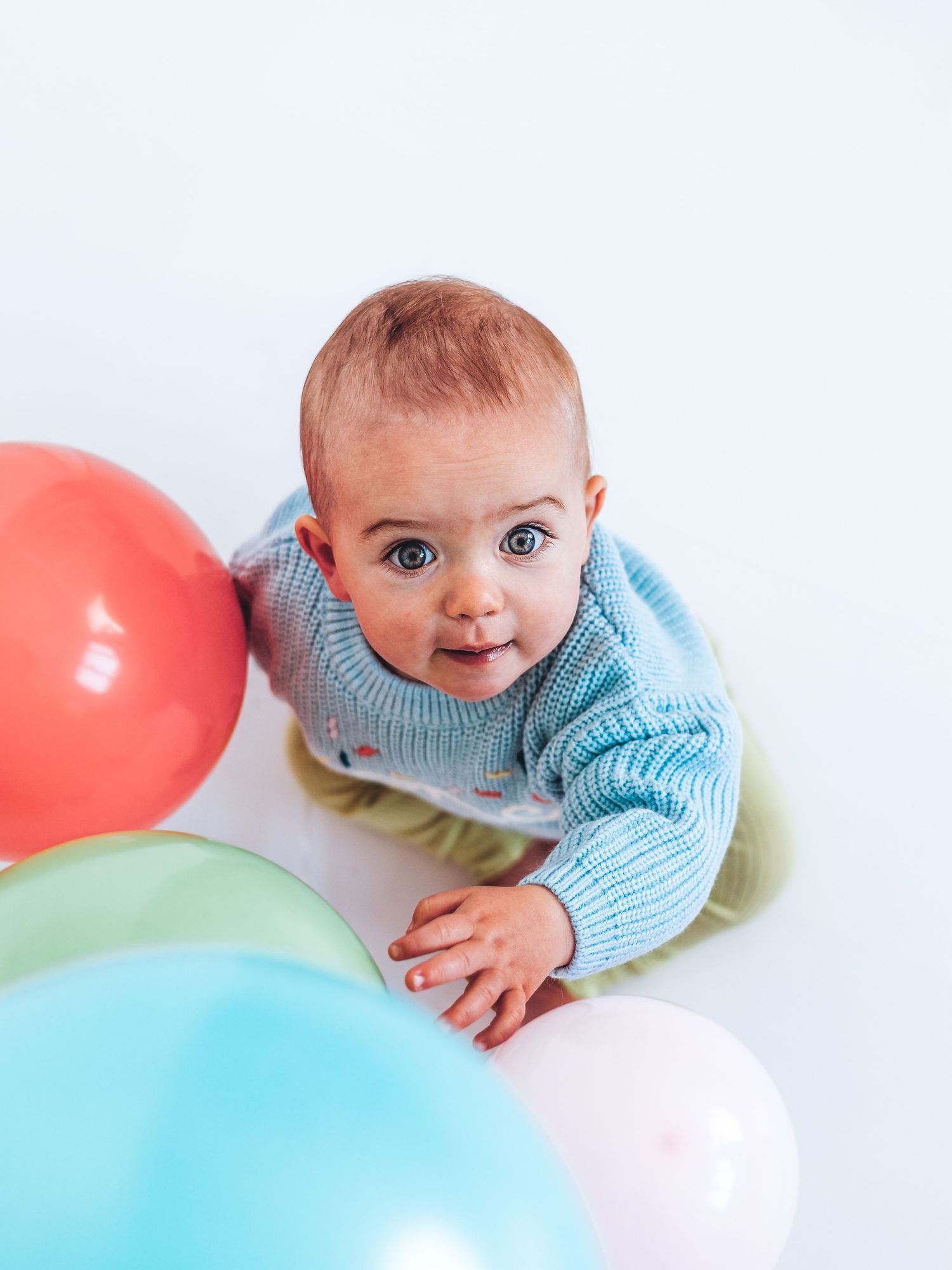 A curious baby in a blue knit sweater surrounded by colorful balloons looks up at the camera on a white background.