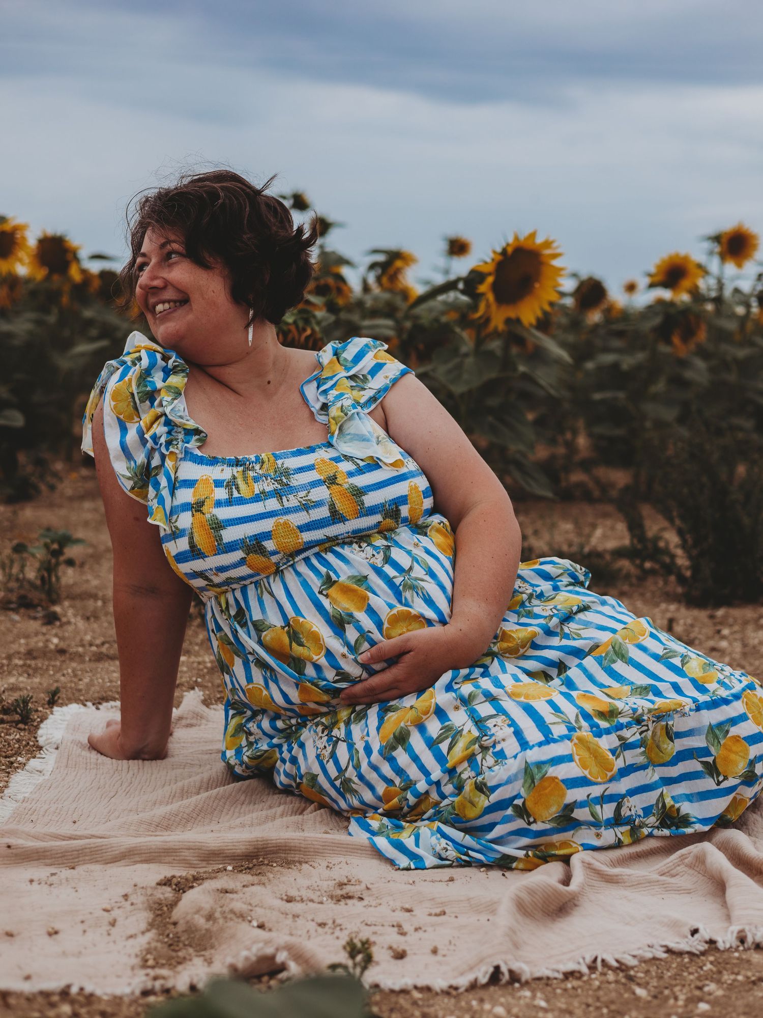 A woman in a blue and yellow sunflower print dress poses playfully on a sandy beach at sunset.