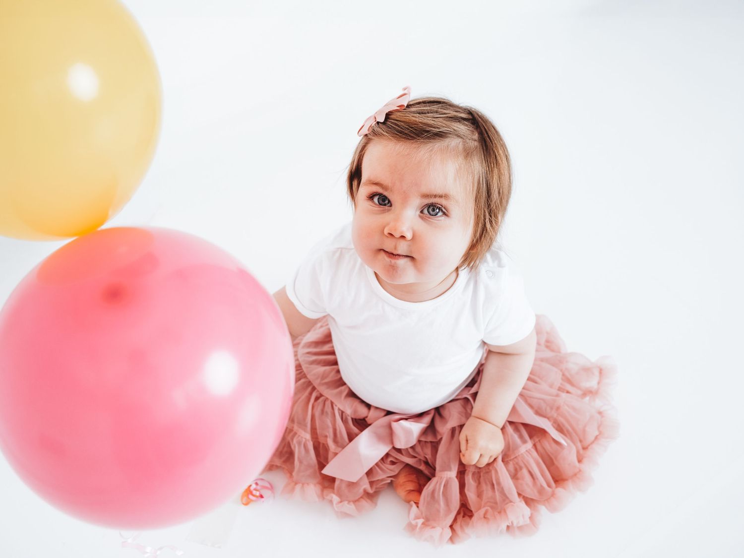 Baby girl in pink tutu skirt sits surrounded by colorful balloons on white background.