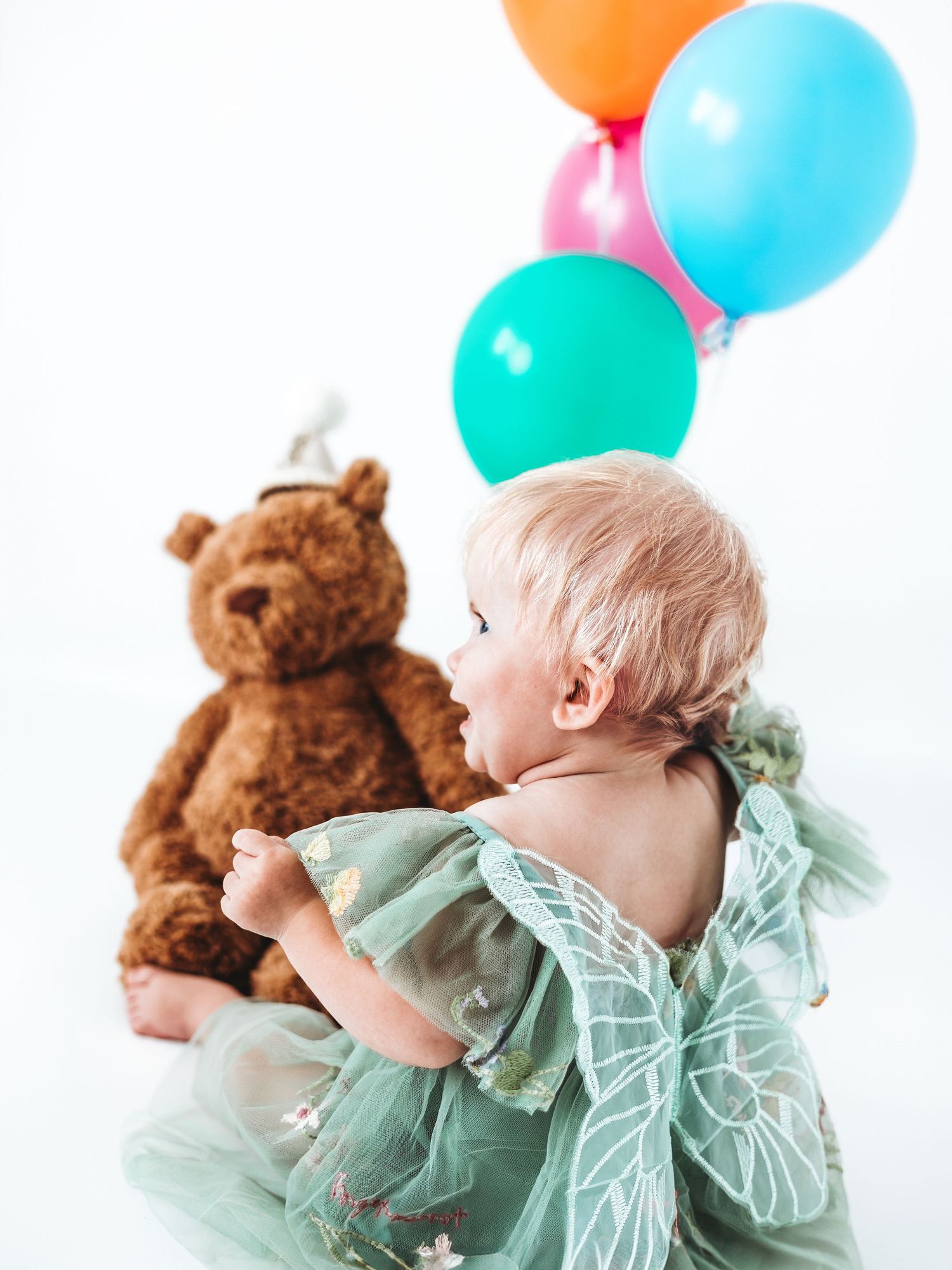 A baby in a green butterfly dress sits beside a teddy bear with colorful balloons on a white background.