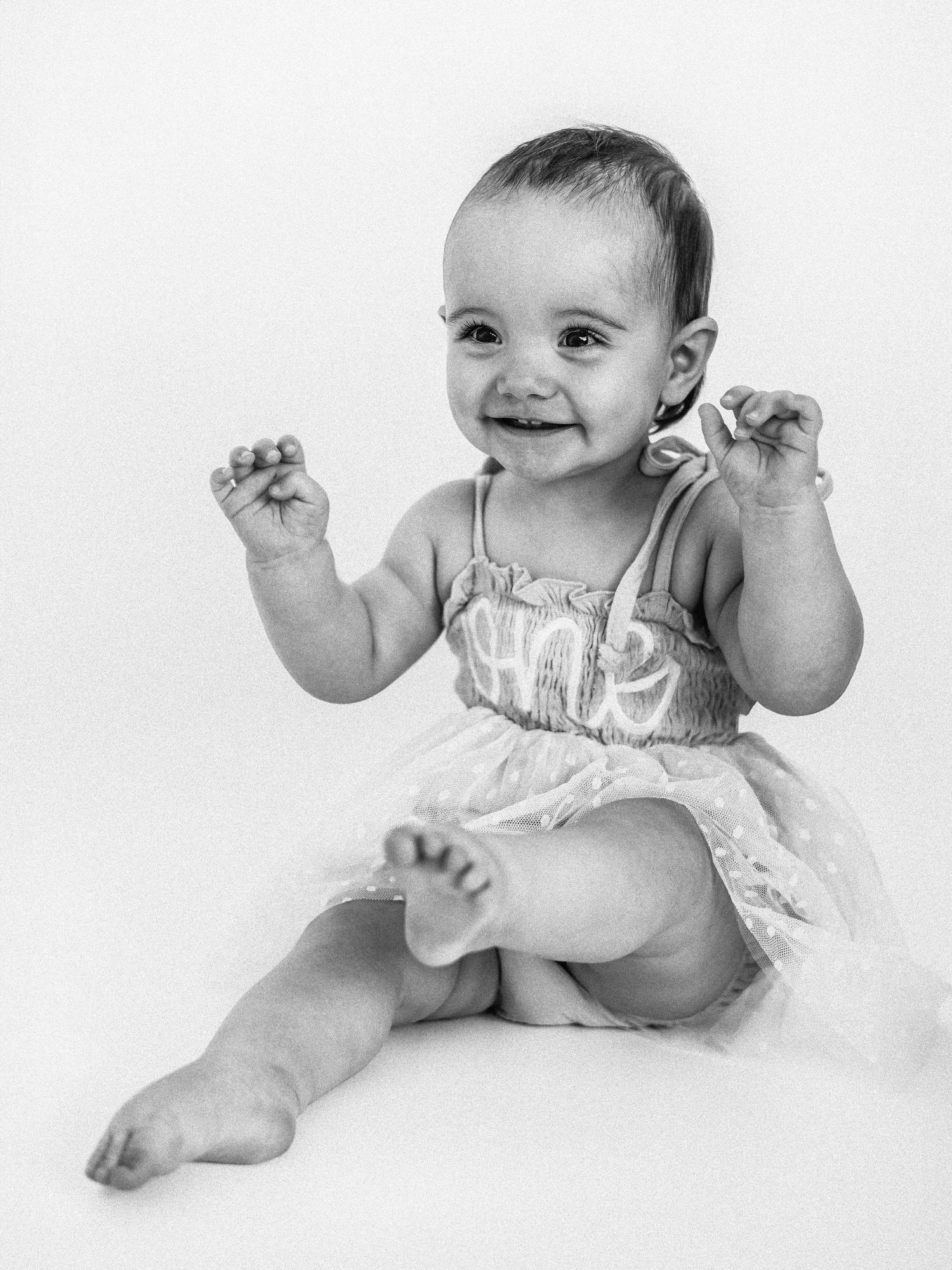 Smiling baby girl in a tutu dress sits upright, raising her tiny fists with joy in a black and white portrait.