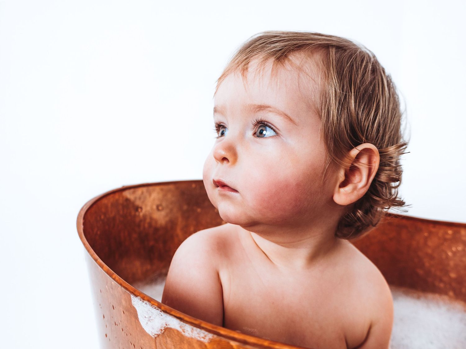 Cute toddler with blue eyes sitting in a rustic copper bathtub with soap suds, looking up curiously.