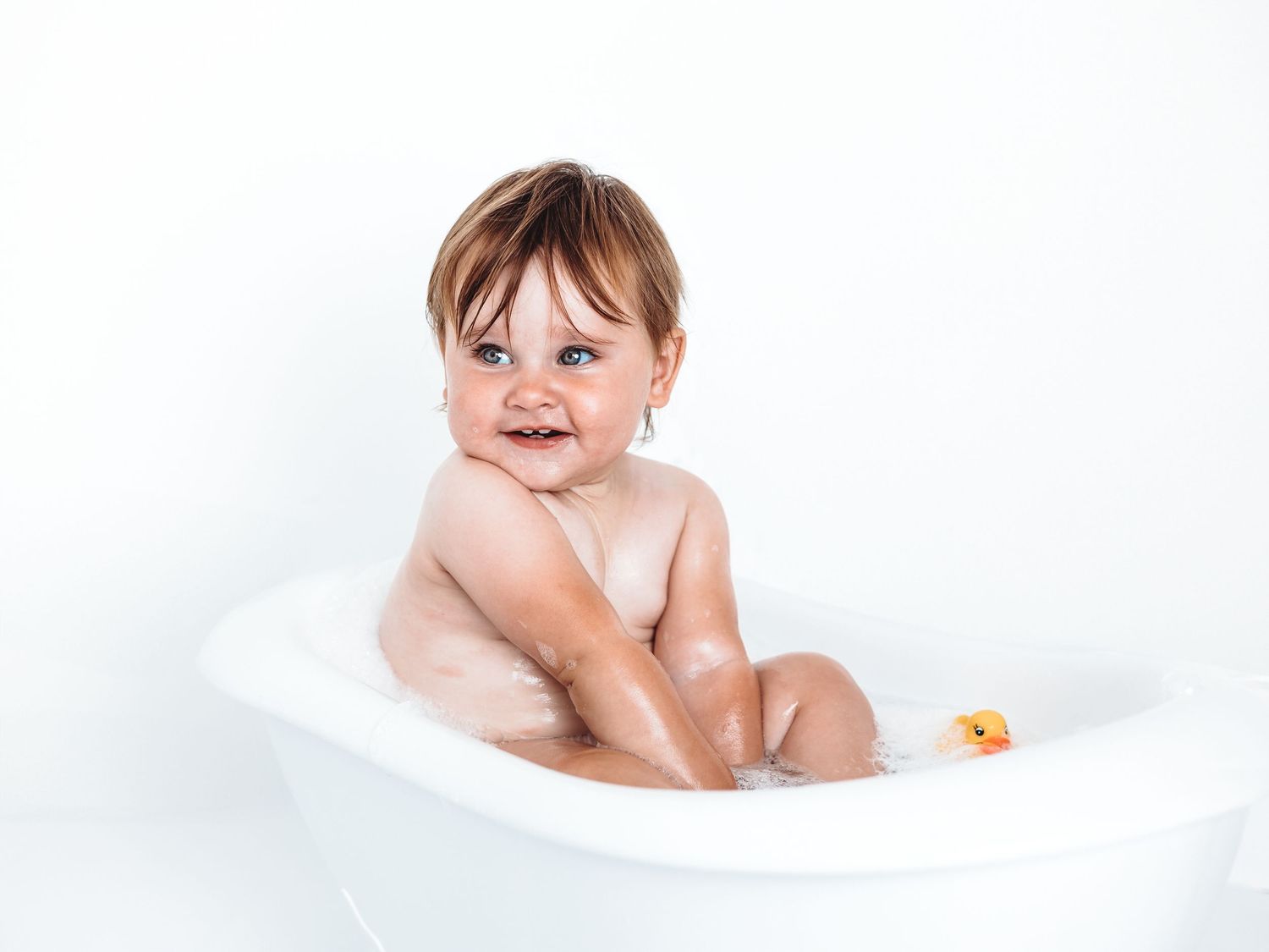 Smiling baby sitting in a white bathtub with bubbles and a rubber duck, enjoying bath time.