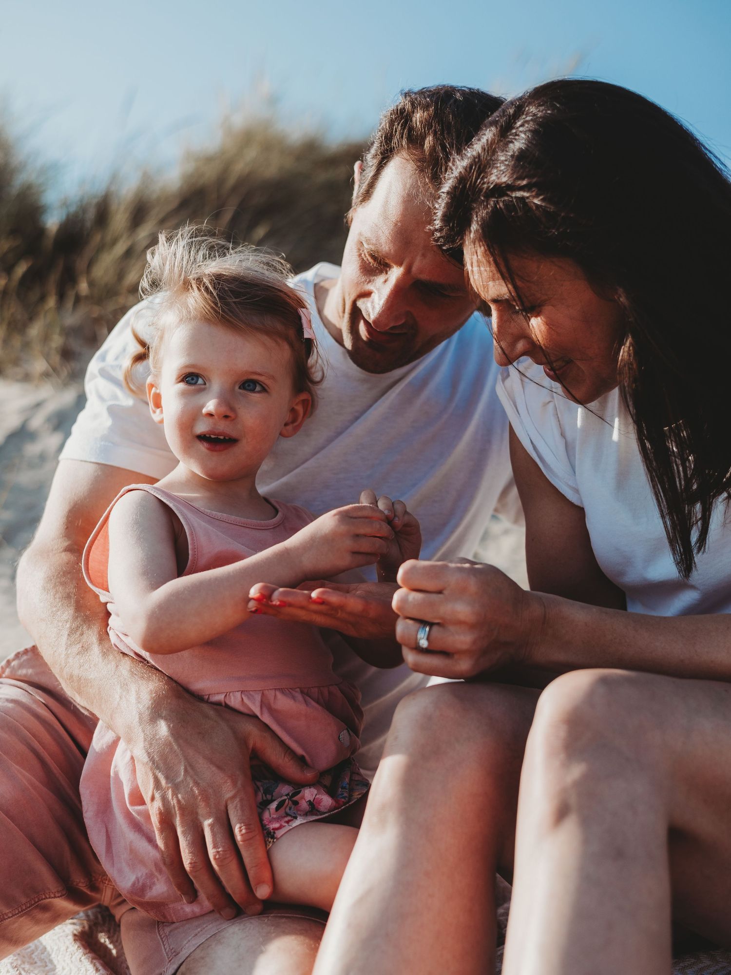 Happy family with toddler girl sitting on sandy beach, parents smiling at child on sunny day.