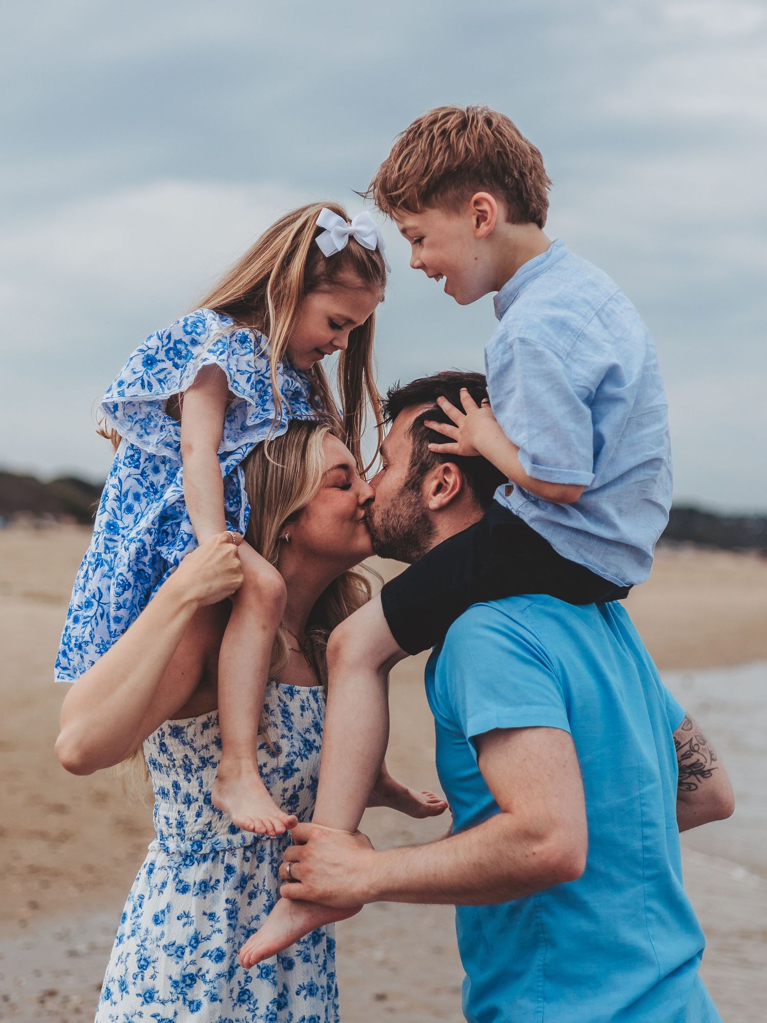 Family of four on beach, parents kissing while lifting young children on shoulders, wearing blue outfits.