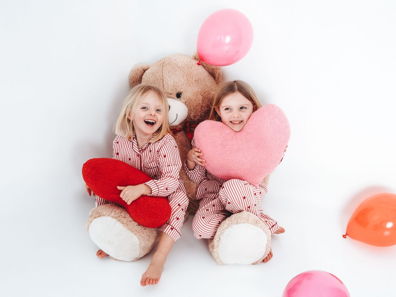 Two young girls in pajamas sitting with a large teddy bear, holding heart pillows and pink balloons on white background.