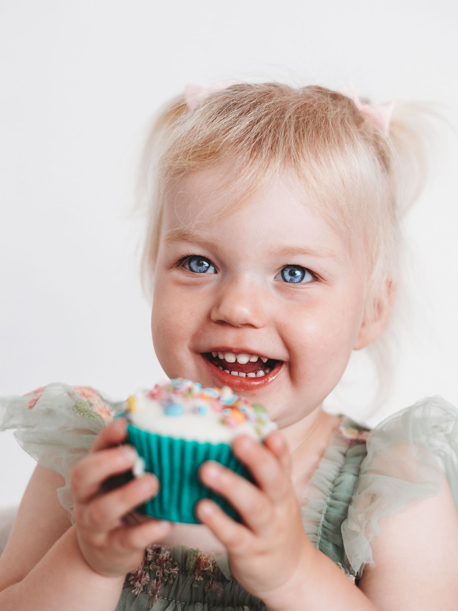 Smiling toddler girl with blonde pigtails and blue eyes holding a colorful sprinkle cupcake in a teal wrapper.
