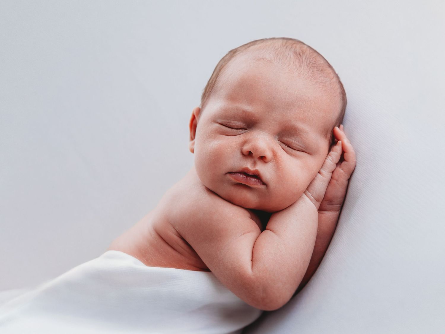 Newborn baby sleeping peacefully on white blanket with gentle hand support.
