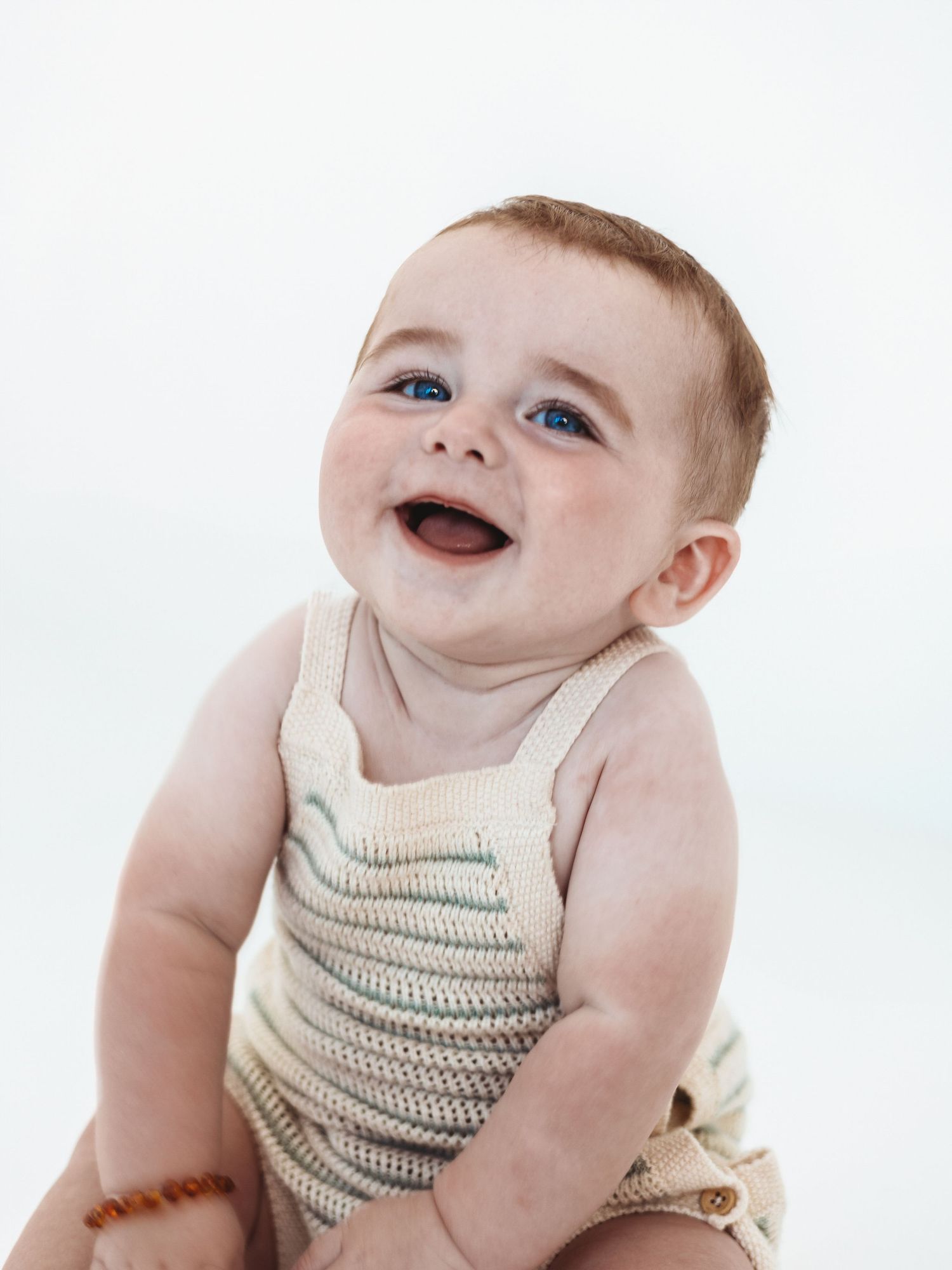A happy baby wearing a cream-colored knit romper smiles while sitting against a white background.