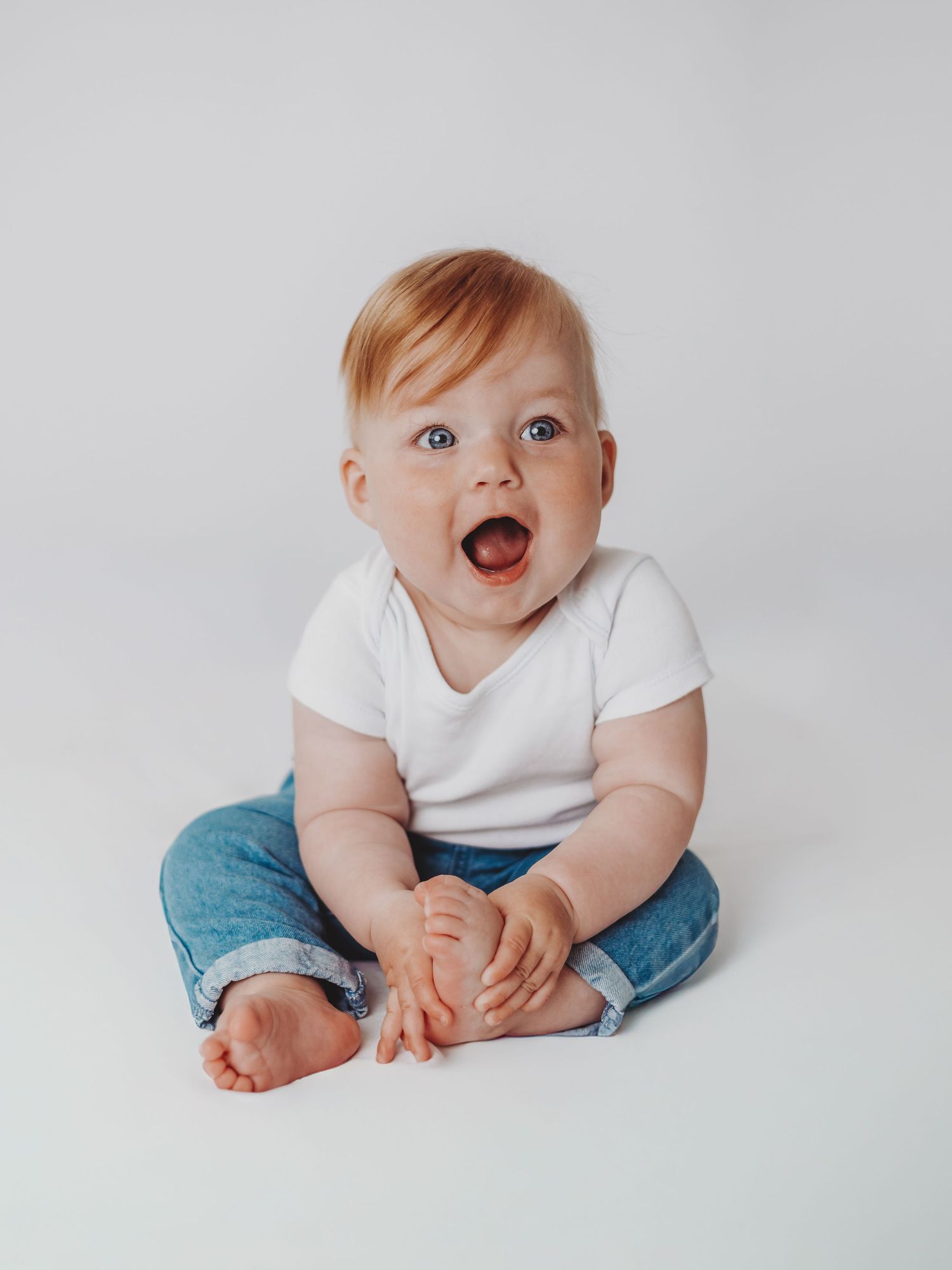 A baby in white shirt and jeans sits on a light gray background in a series of candid poses showing playful expressions.