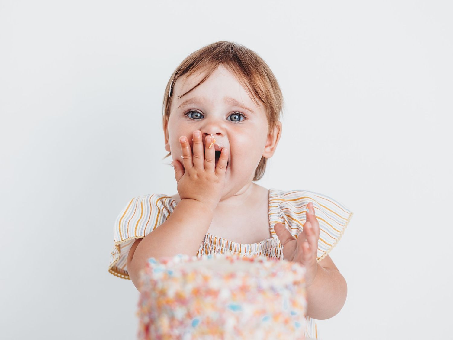A baby in a polka dot dress with ruffled sleeves laughs while covering their face in a series of playful expressions.