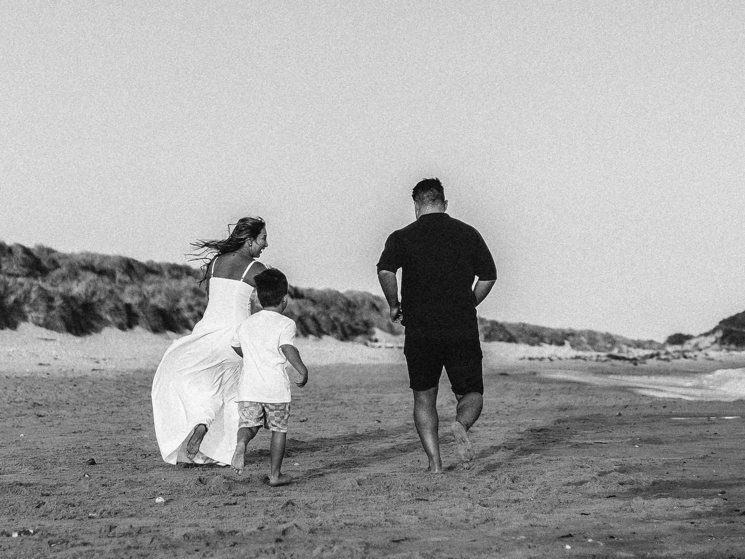 Black and white photo of a family walking on a sandy beach, with a woman in a white dress, a child, and a man in black.