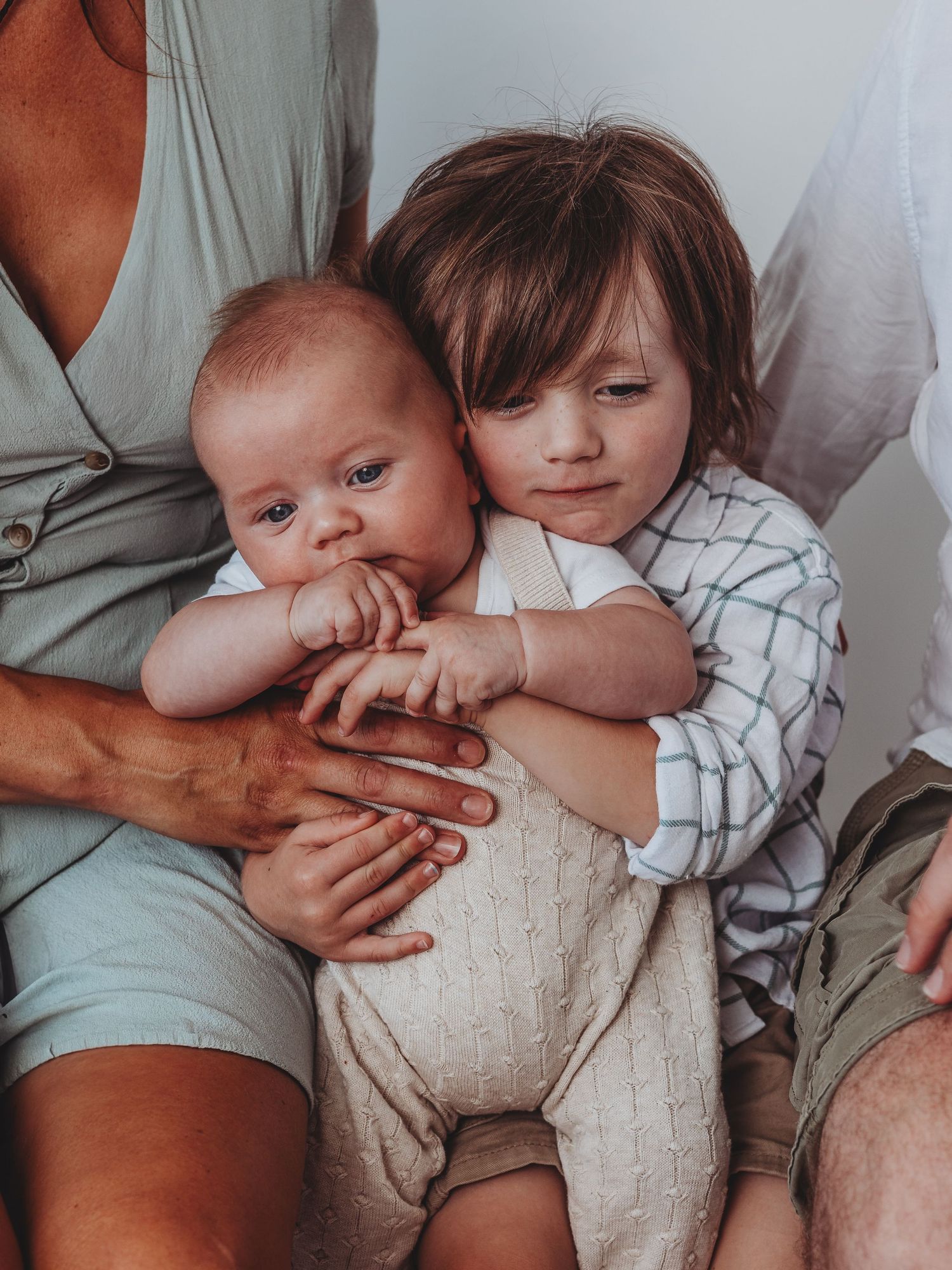 A toddler gently hugs a newborn baby while sitting on a parent's lap in a warm, candid family portrait.