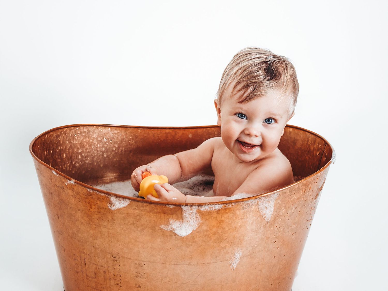 Happy baby sitting in a rustic copper bathtub holding a yellow toy duck, splashing in soapy water.