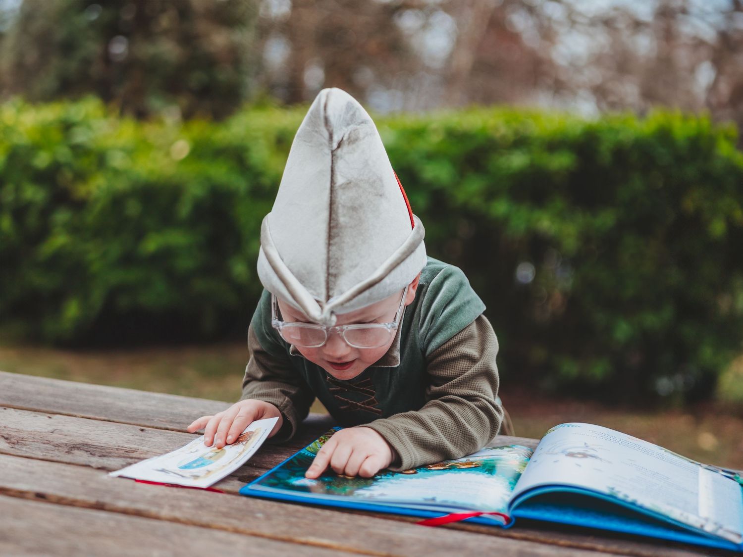 Young child wearing a pointed gnome hat and glasses reads a colorful picture book outdoors at a wooden table.