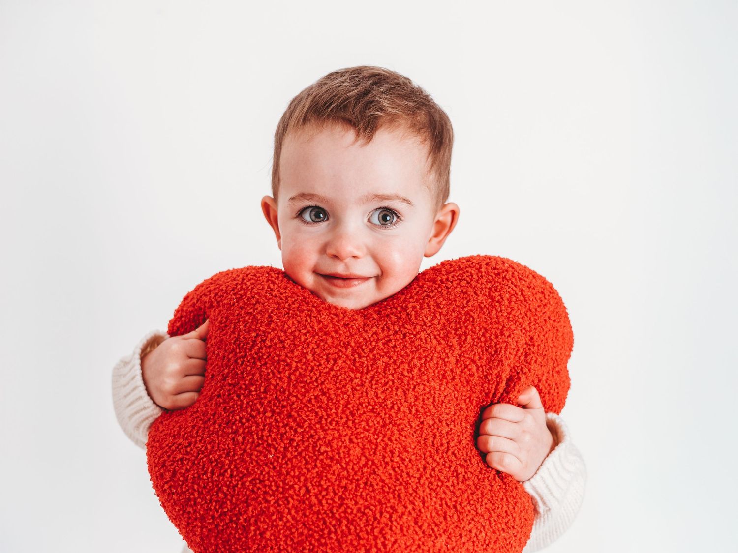Adorable baby with blue eyes hugging a red heart-shaped pillow against a white background.