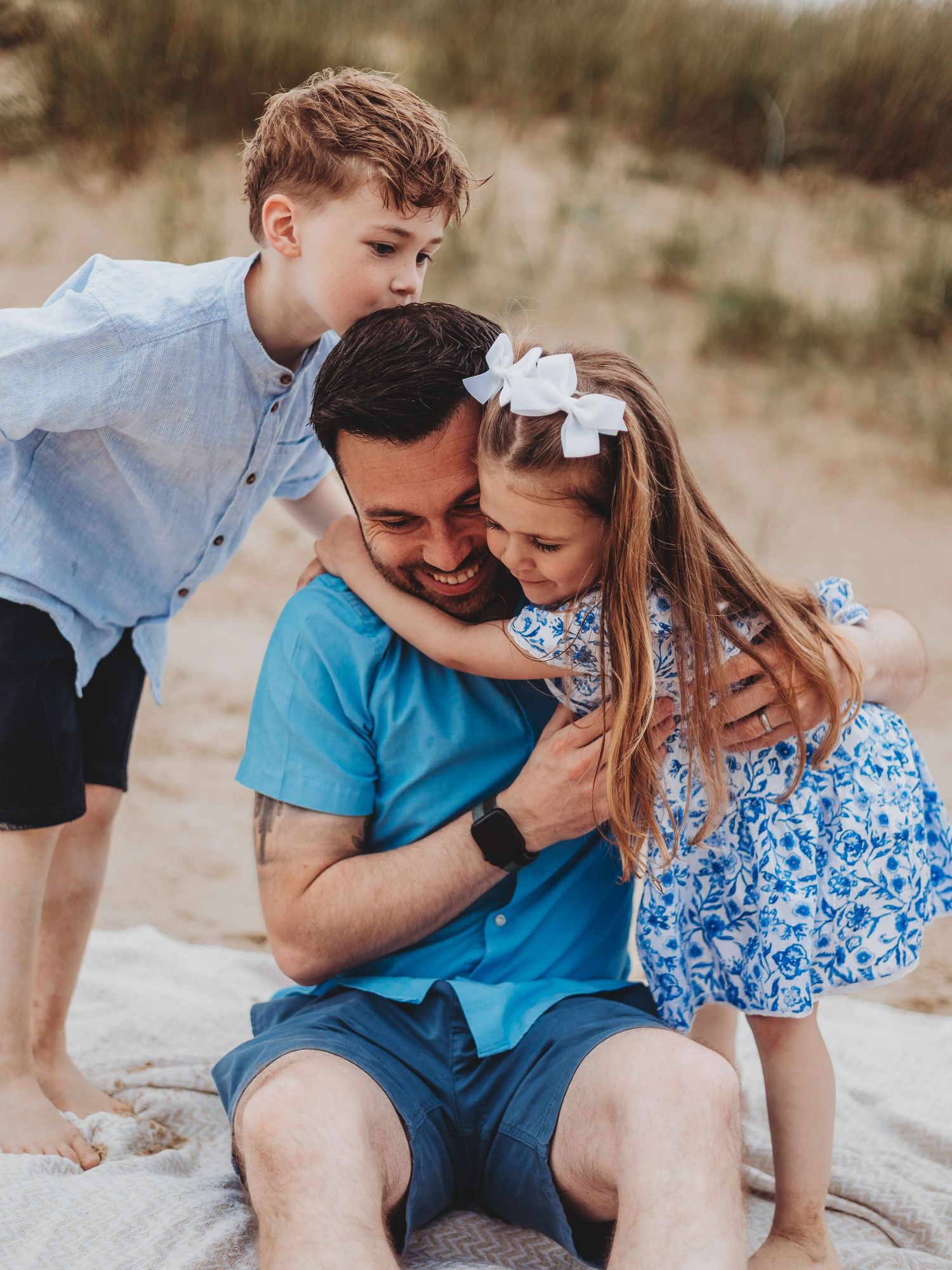 A father sits on a sandy beach hugging his two young children, a boy and girl, in a heartwarming family moment.
