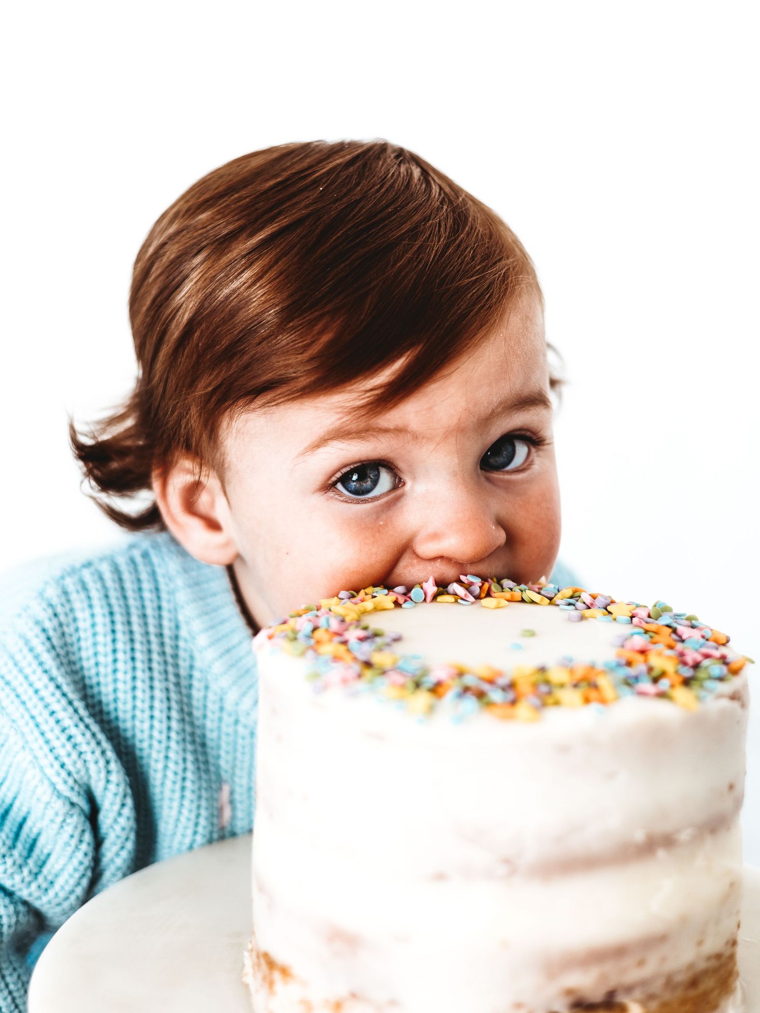 Cute toddler in blue sweater biting into a white birthday cake topped with colorful sprinkles.