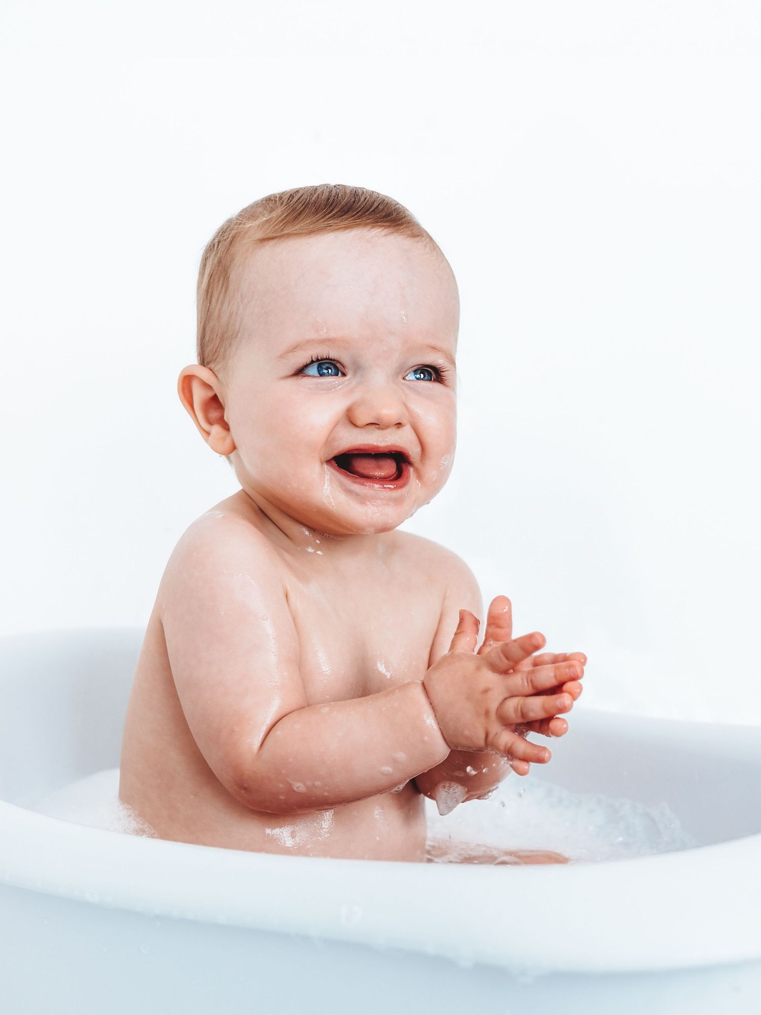 Happy baby with blue eyes clapping hands while sitting in a white bathtub with bubbles.