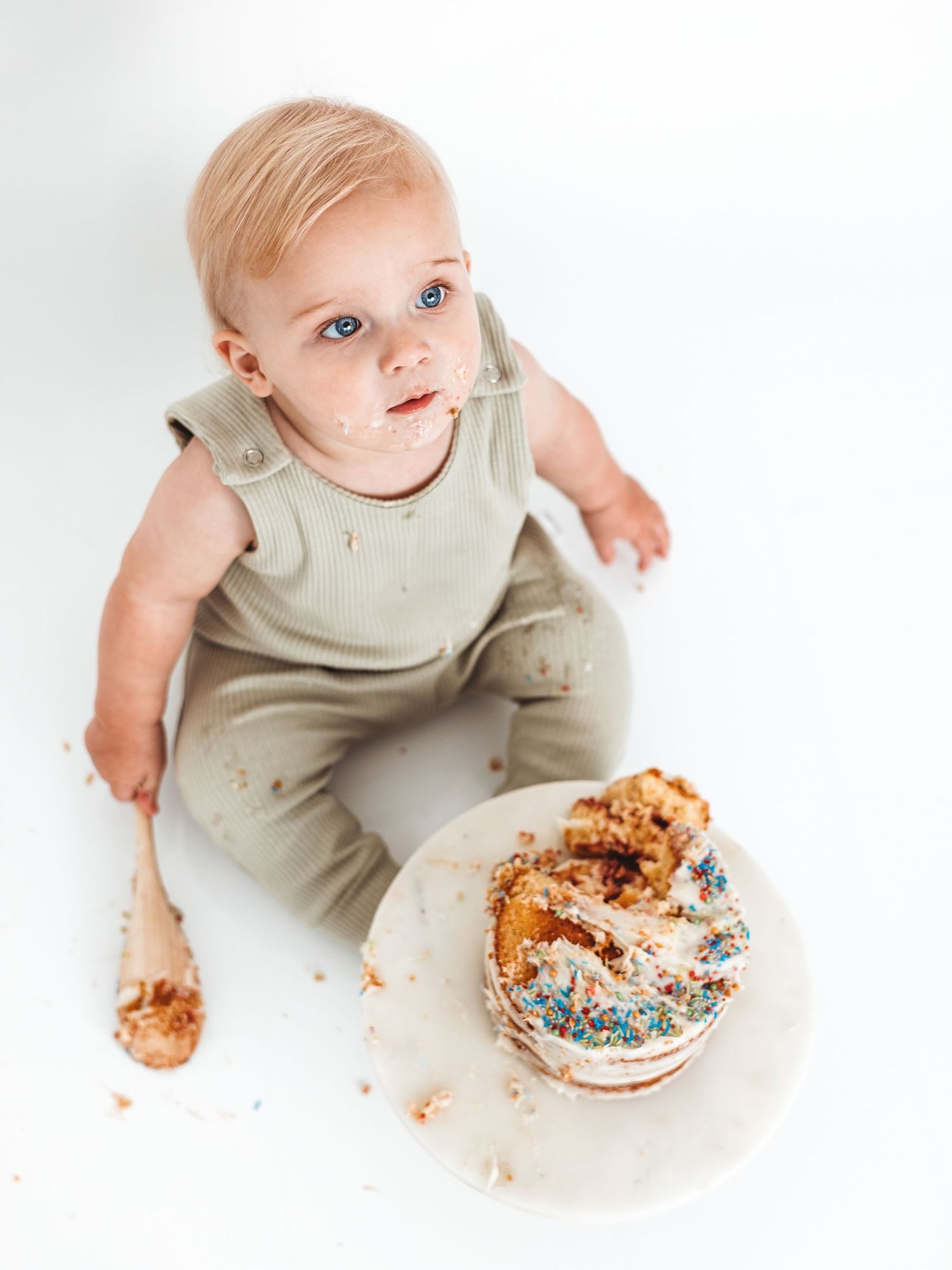 Baby in sage romper sits with smashed first birthday cake and wooden spoon on white background.