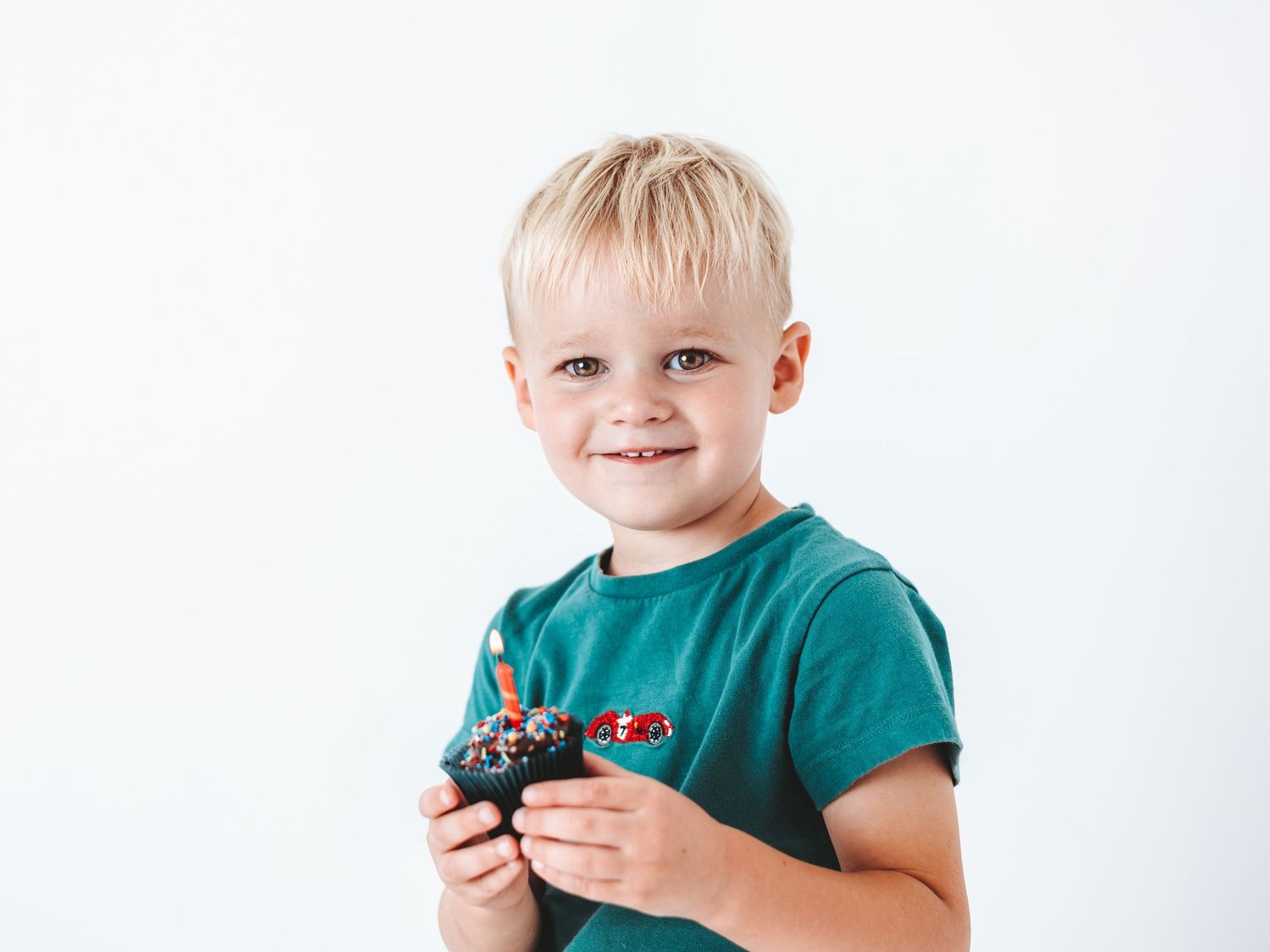 Smiling blonde toddler boy in green t-shirt holding a chocolate cupcake against a white background.