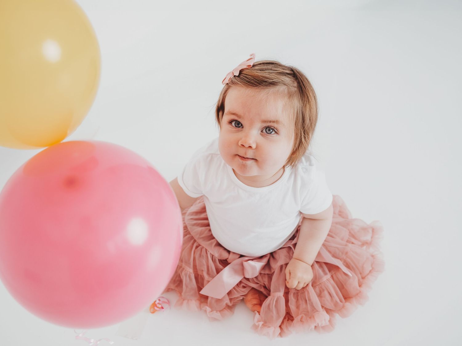 A baby in a pink tulle dress sits on a white backdrop with pink and yellow balloons in a studio setting.