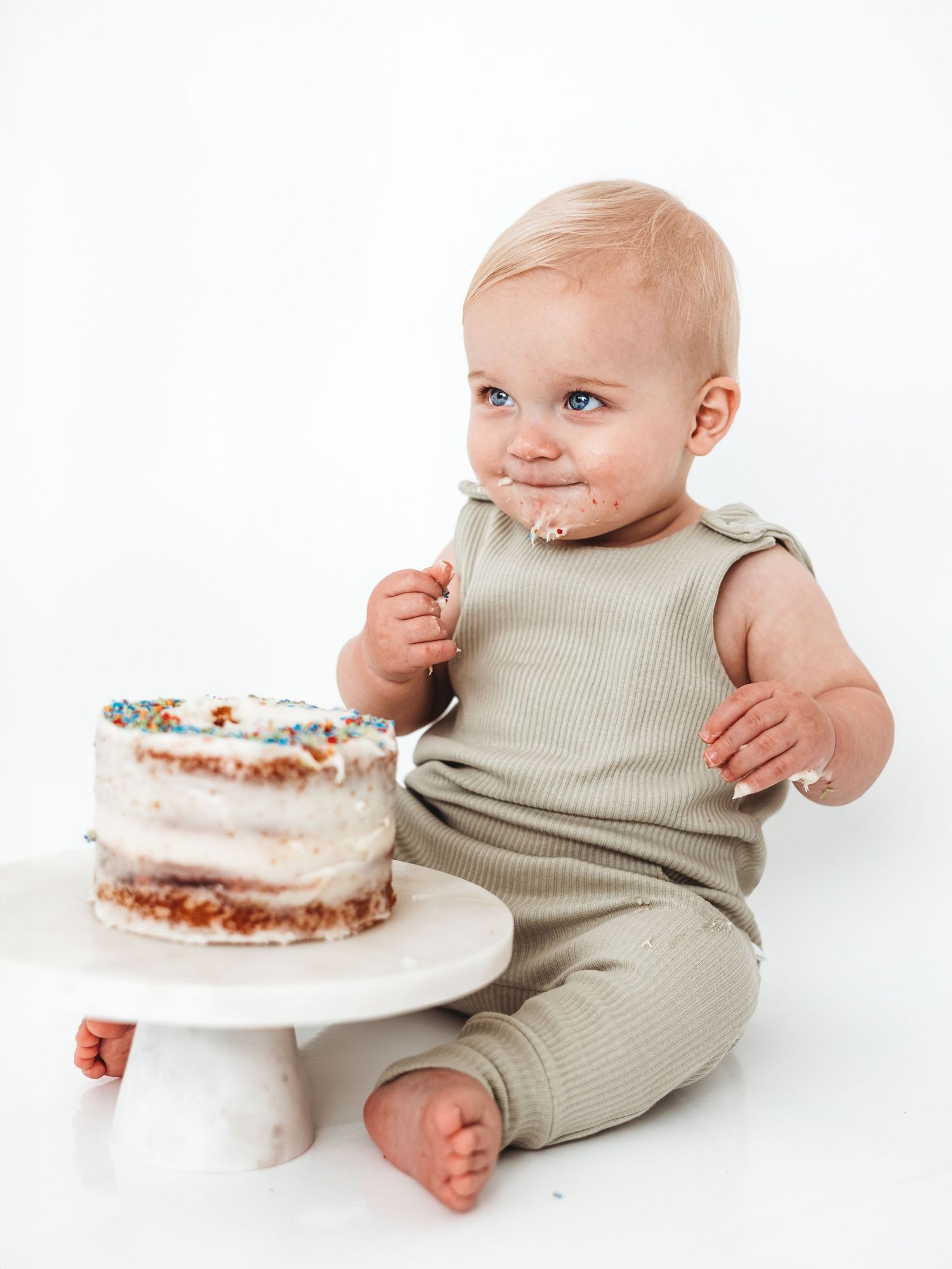 Baby in sage green romper smashing first birthday cake with sprinkles on white background.