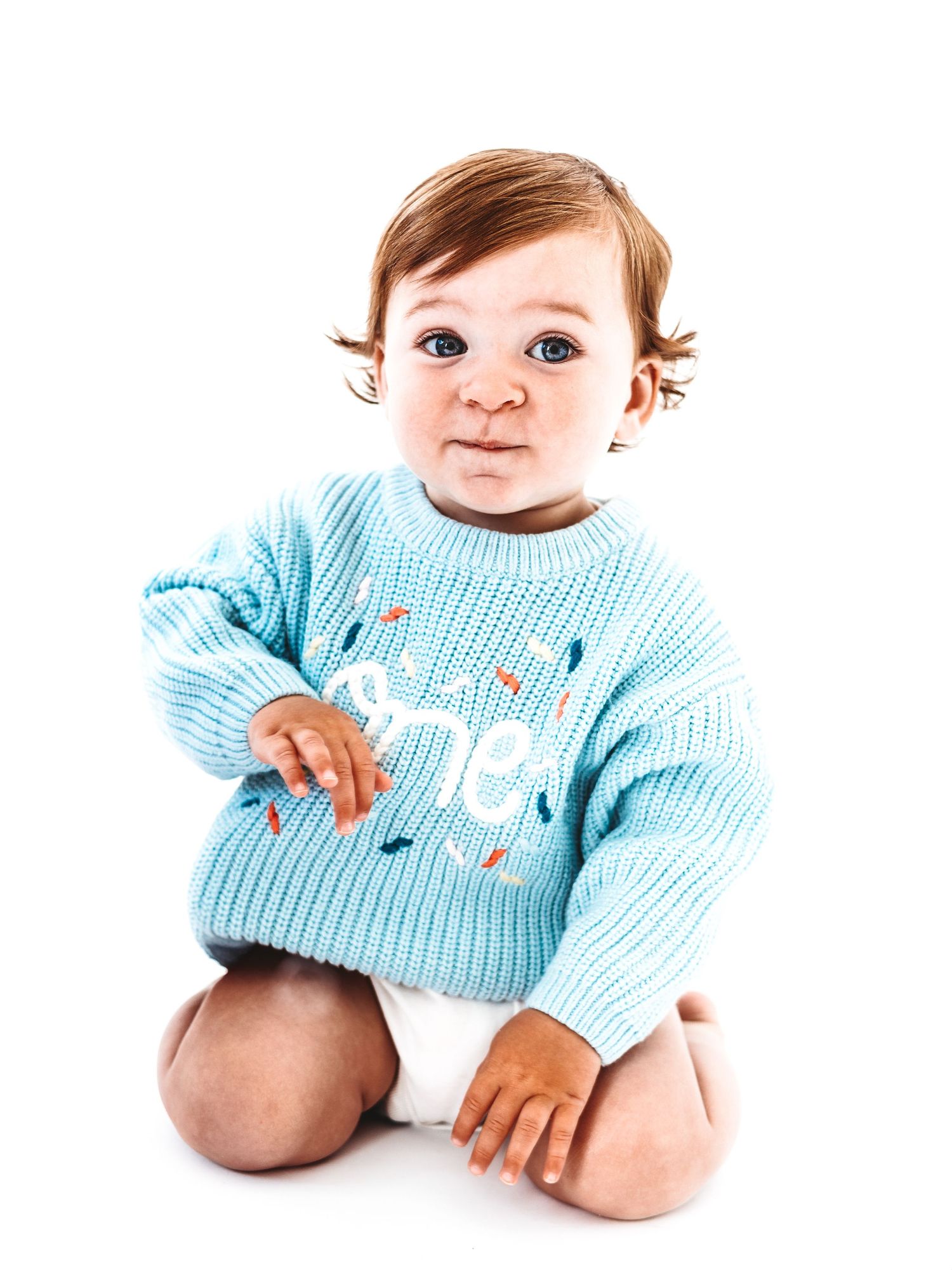 Baby wearing a blue knit sweater with 'one' text sits on white background, celebrating their first birthday.
