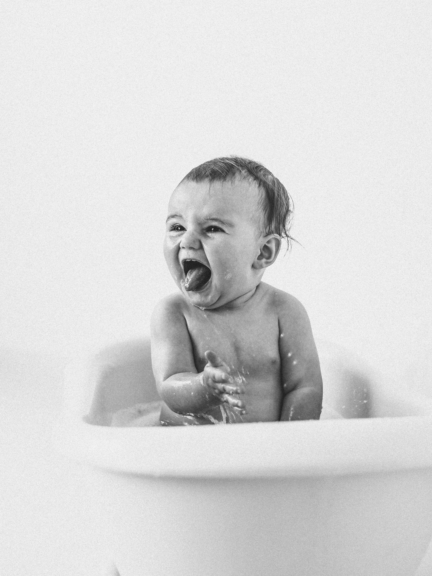 Black and white photo of a happy laughing baby splashing in a small white bathtub.