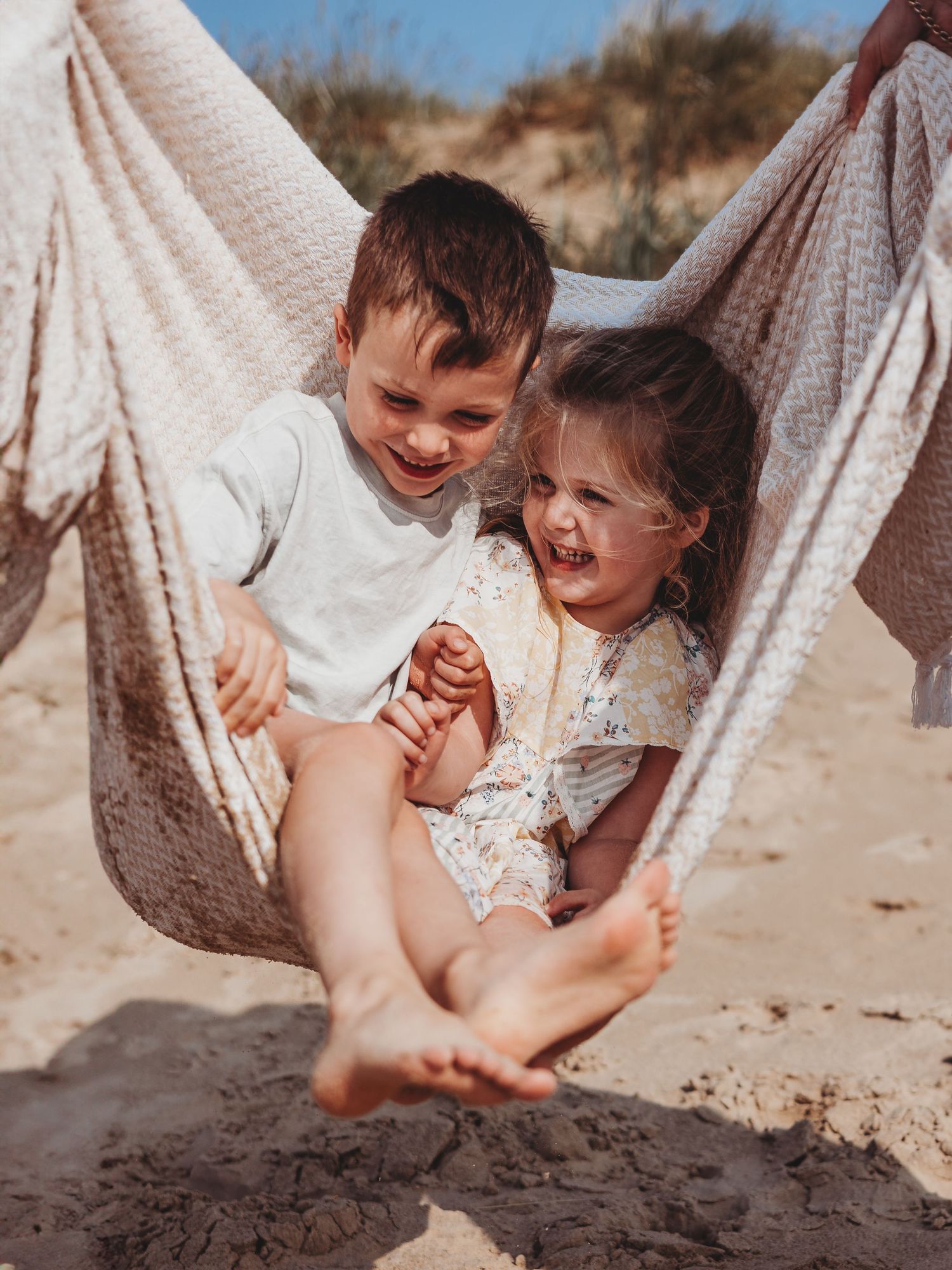 Two happy children laughing and playing together in a hammock on a sunny sandy beach.