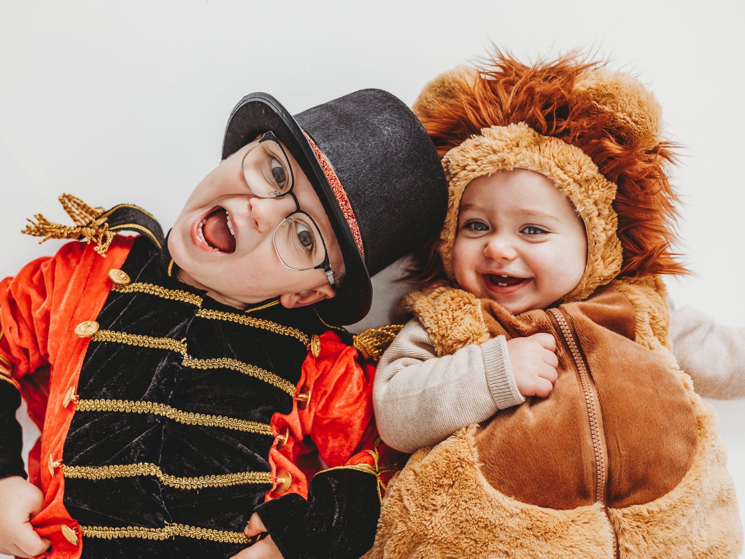 Two children in Halloween costumes, one dressed as a ringmaster with glasses and one as a cute lion, smiling together.