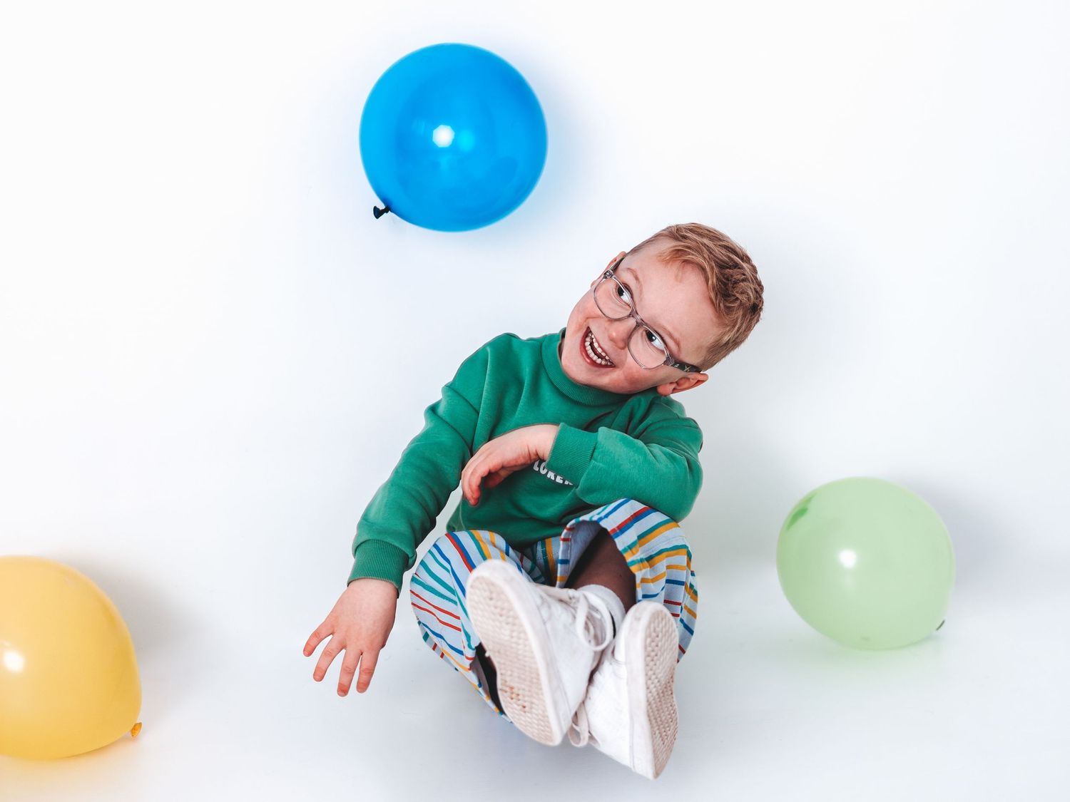 Smiling boy in green sweater and glasses sitting on floor surrounded by colorful balloons on white background.