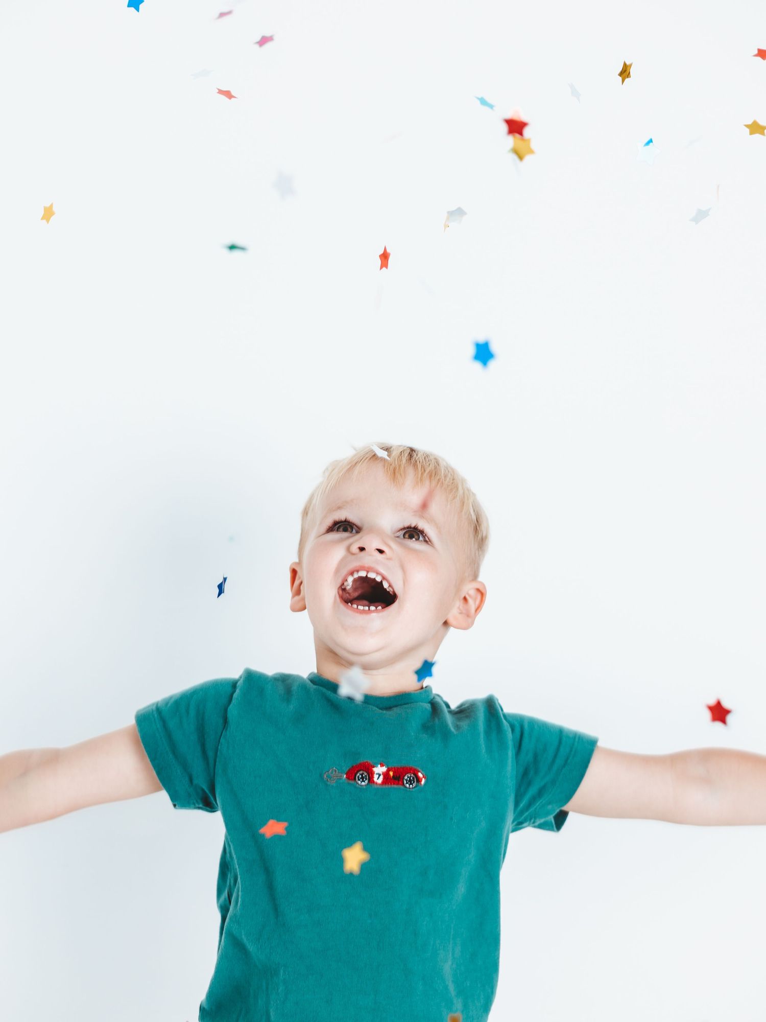 Joyful blonde toddler in green tee catching colorful star confetti with arms outstretched on white background.