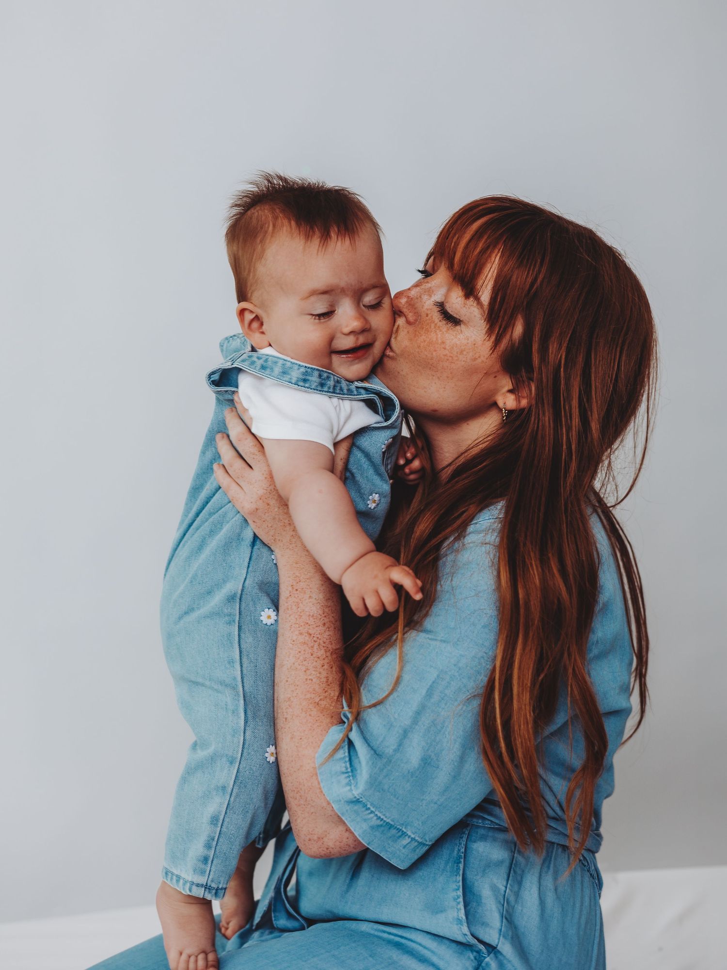 A red-haired mother in denim kisses her smiling baby dressed in matching denim overalls against a white background.