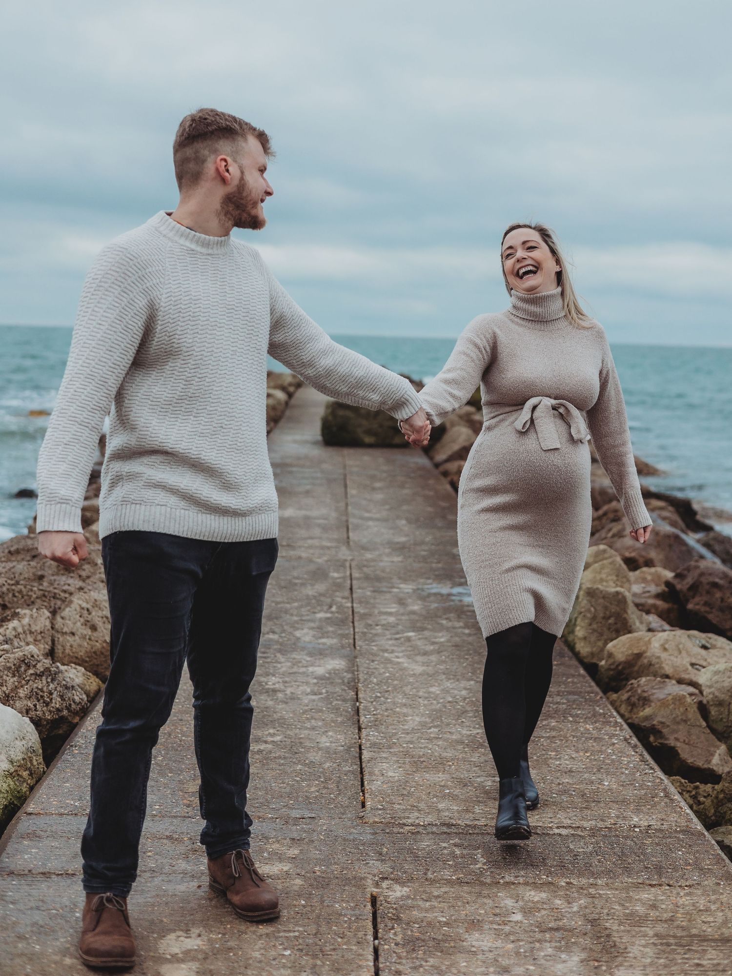 A couple in matching beige sweaters walks along a rocky coastal pier on an overcast day.