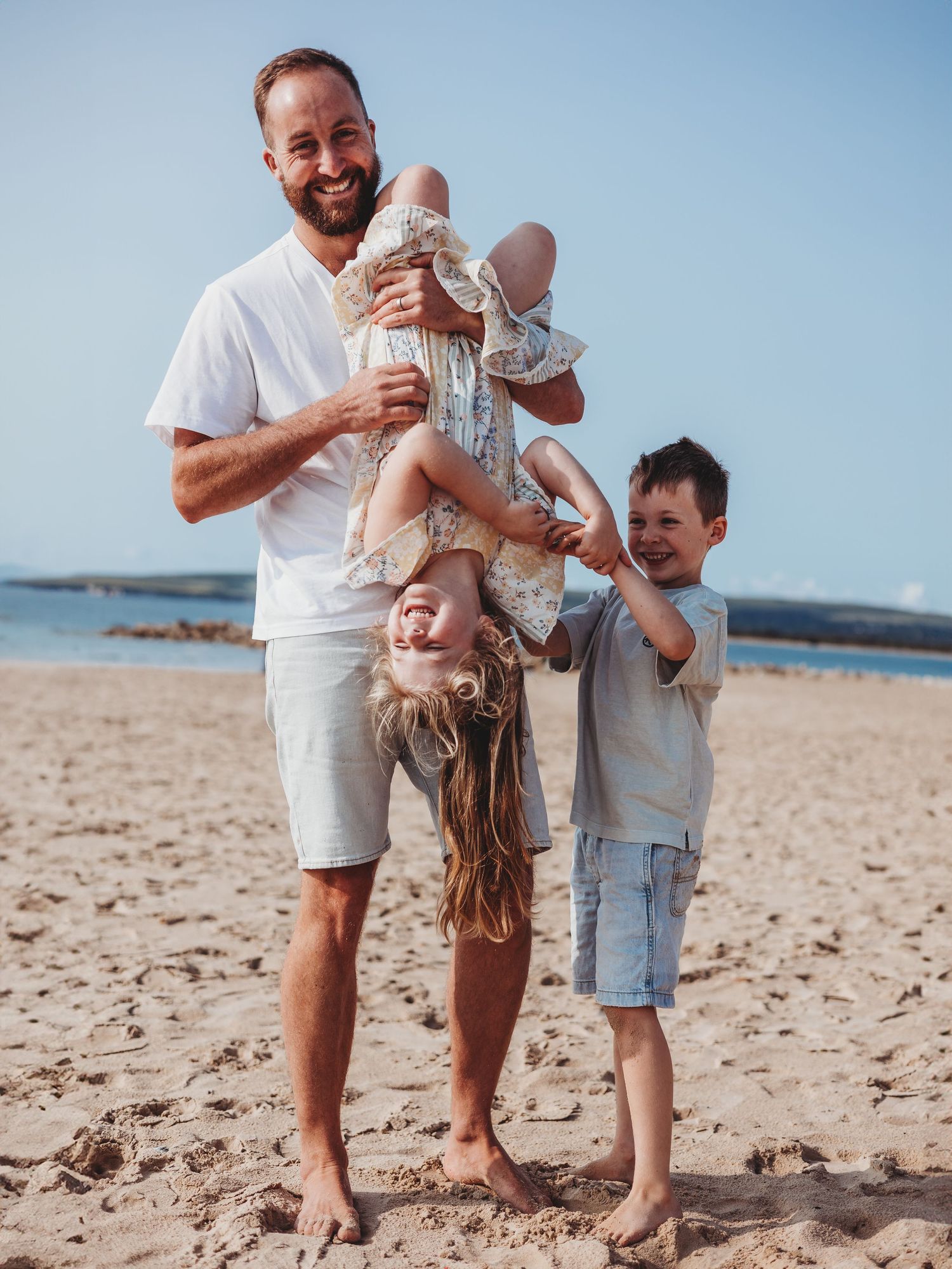 A smiling man holds two laughing children upside down and right-side up on a sunny sandy beach.