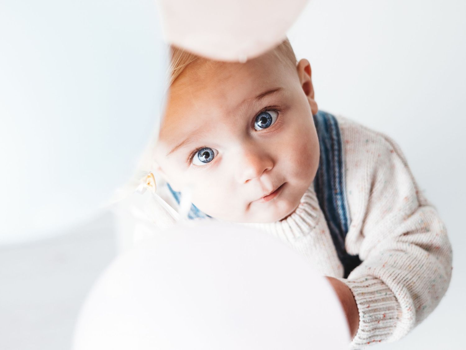 Curious blue-eyed baby in a beige knit sweater peering over a white object against a light background.