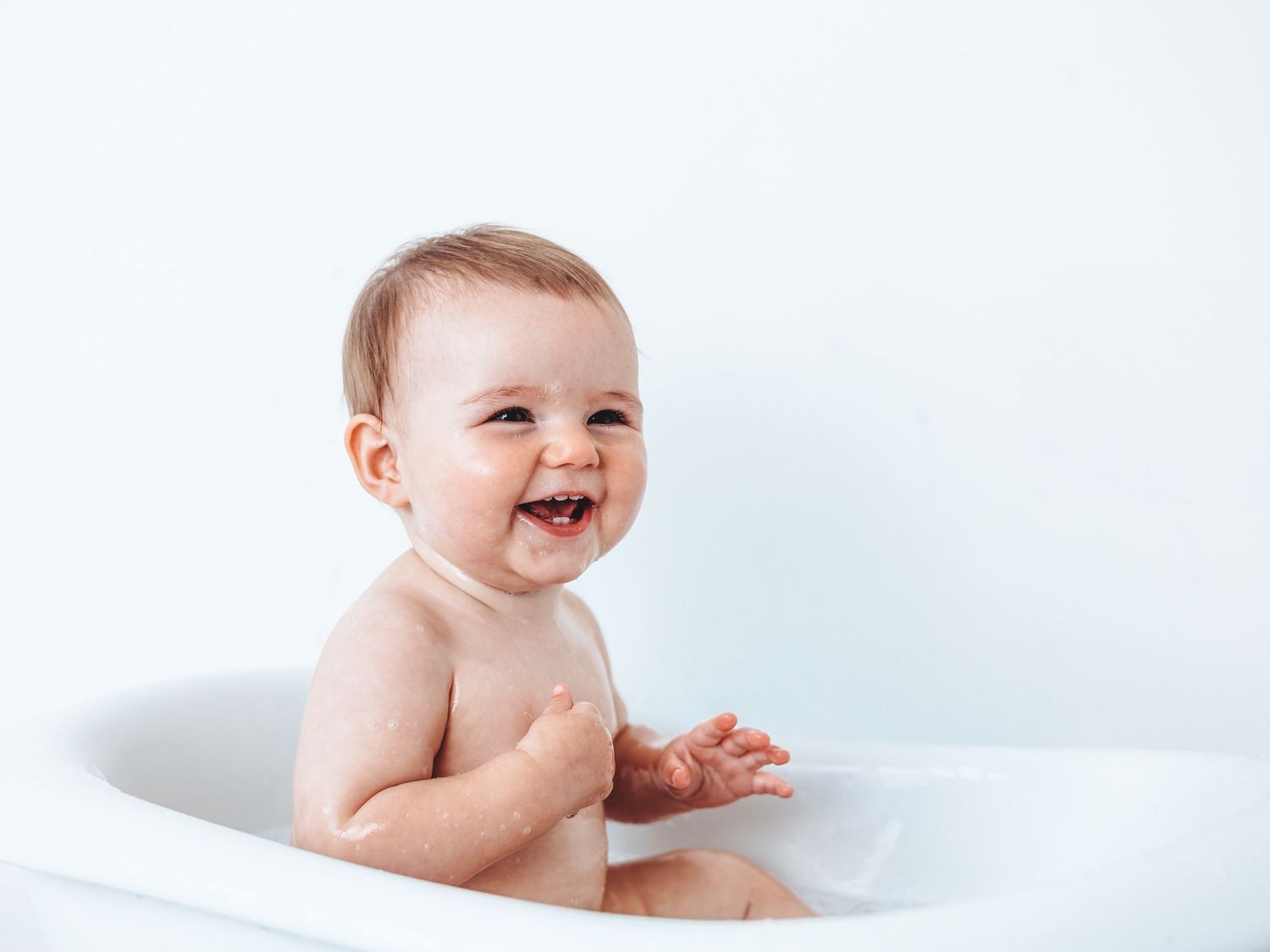 Happy baby laughing in a white bathtub, enjoying bath time in a bright, clean bathroom setting.