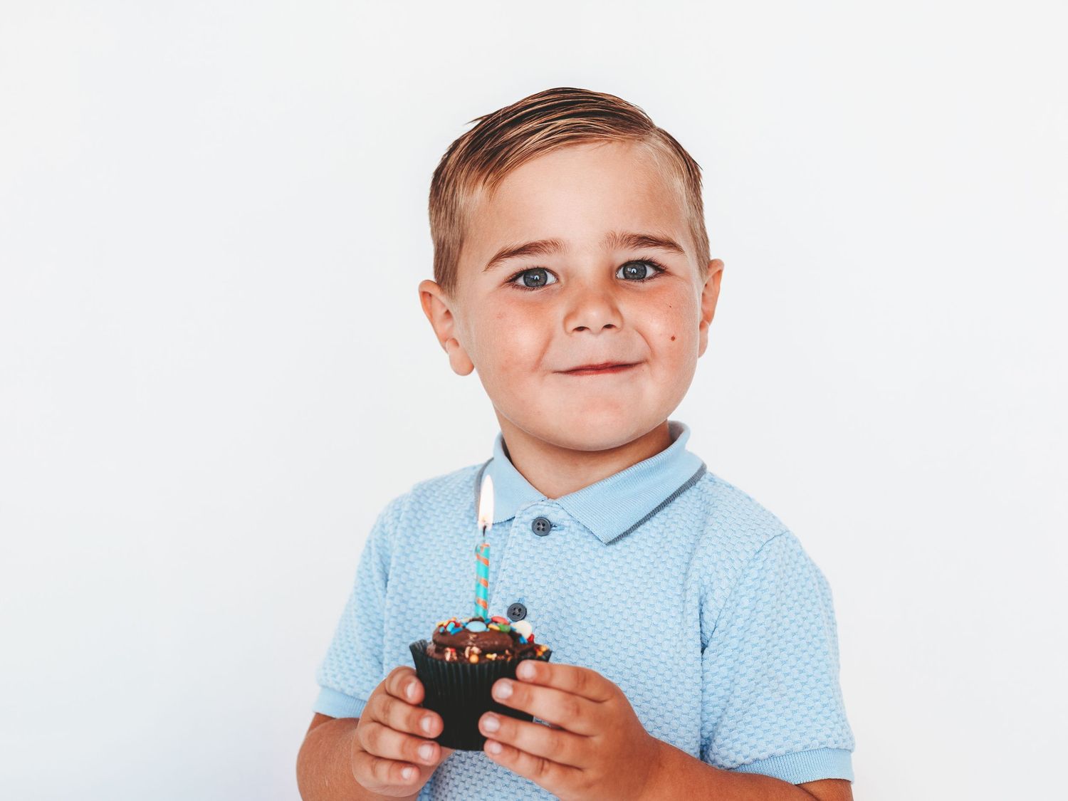 Young boy in blue polo shirt smiling while holding a cupcake with a candle on white background.
