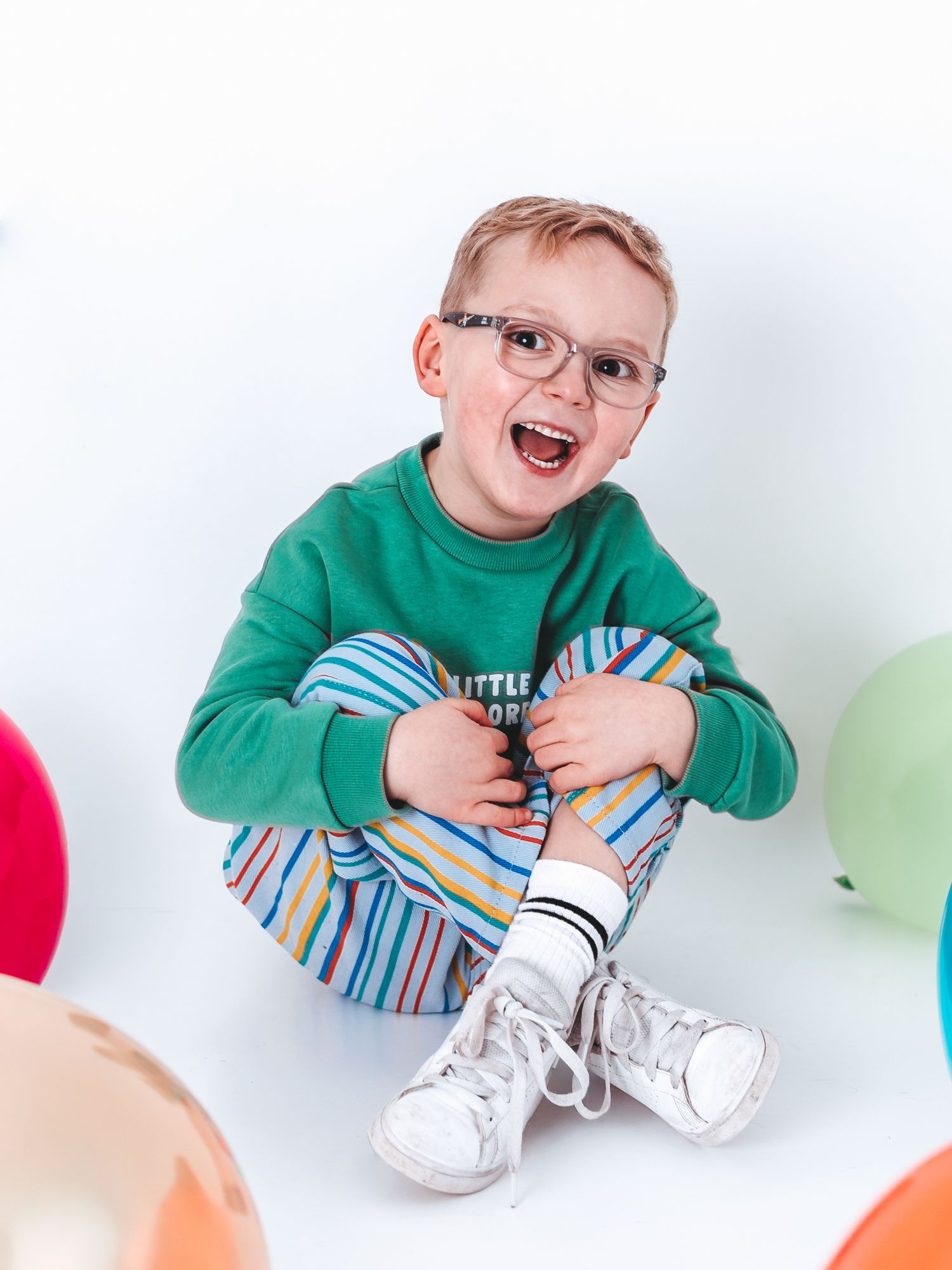 Cheerful young boy with glasses wearing a green sweatshirt and striped pants, sitting cross-legged among colorful balloons.